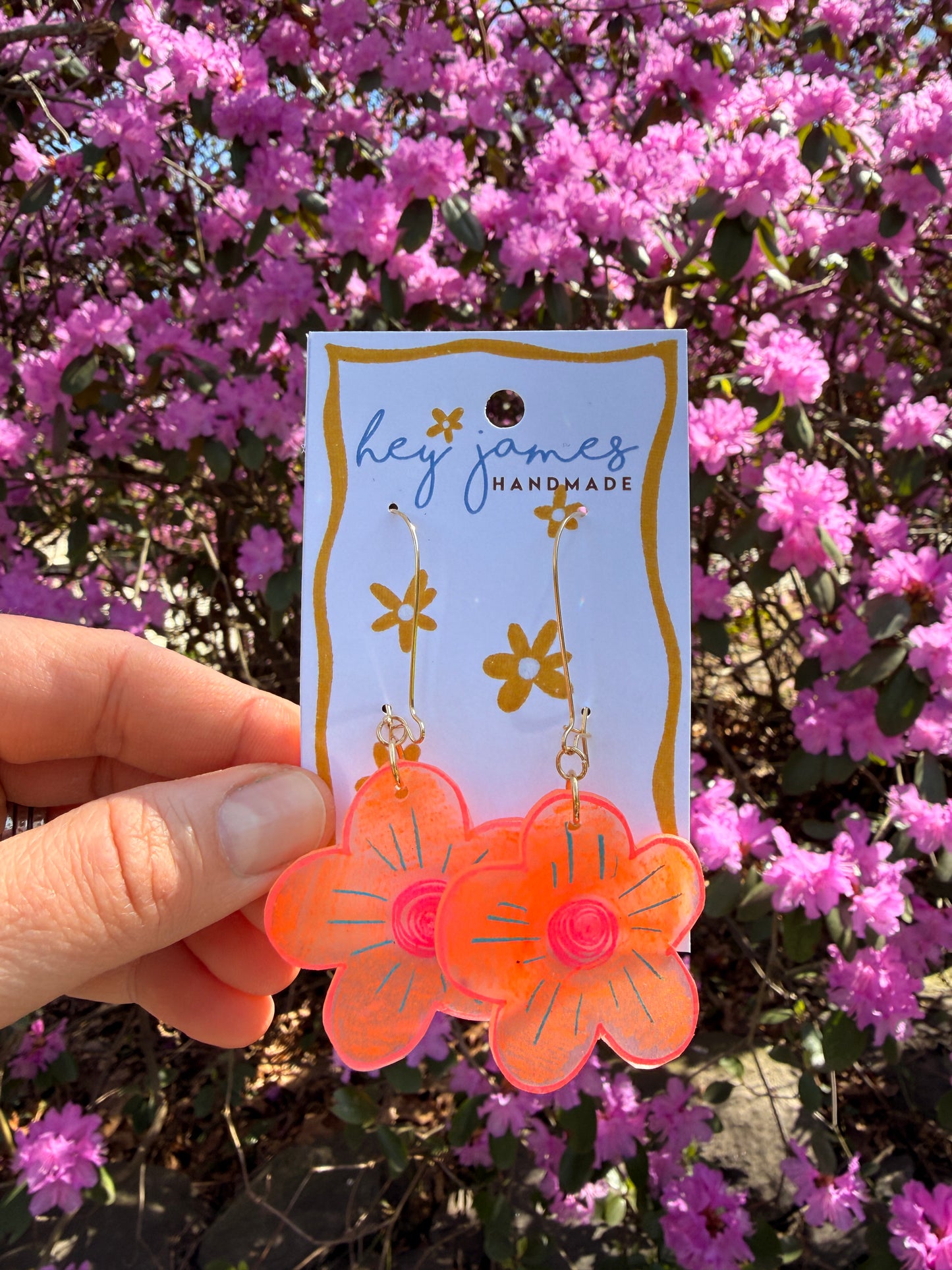 Hand holding floral-themed orange earrings in front of pink flowers