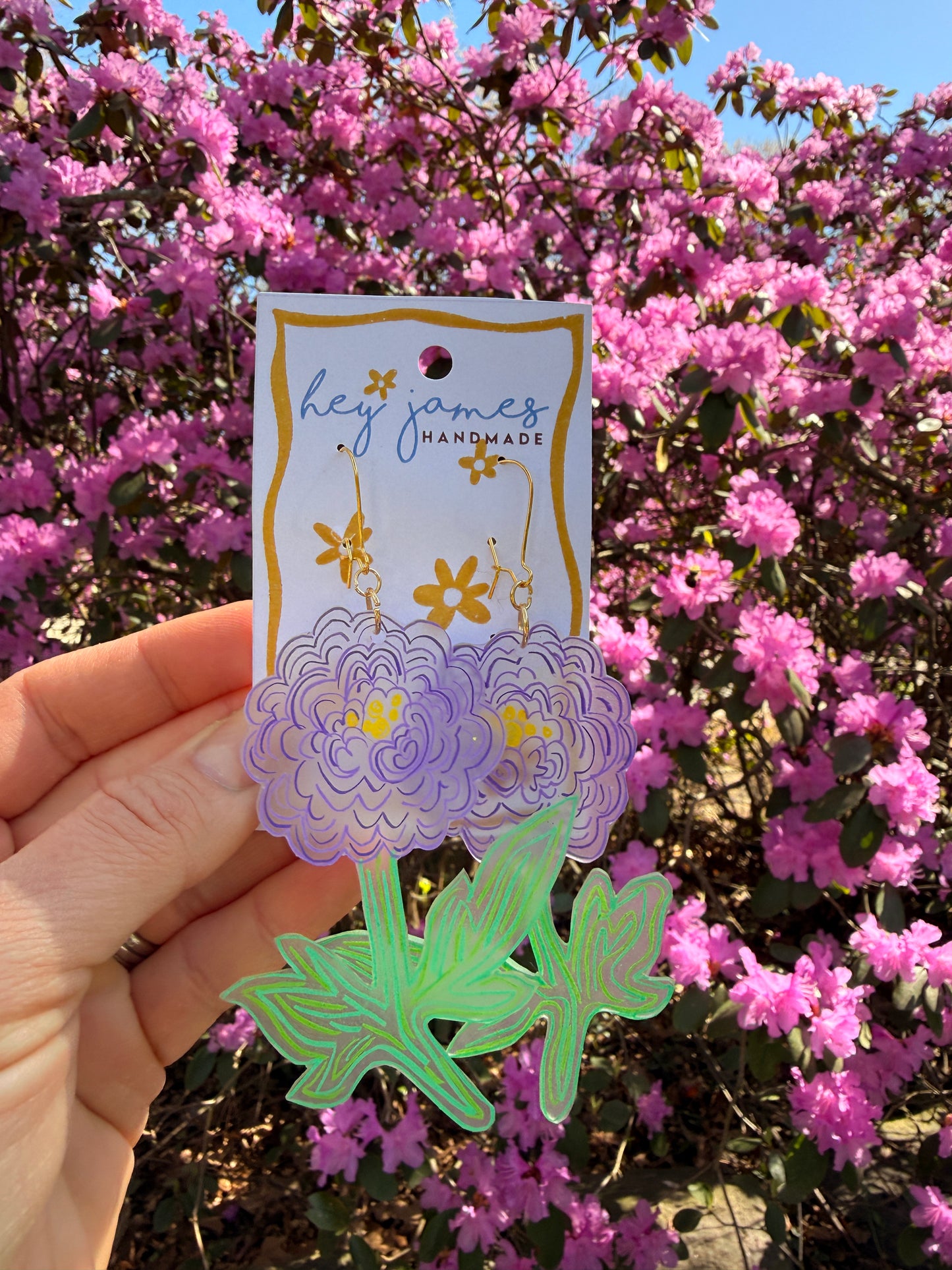 Hand holding decorative purple flower earrings with a floral design against a background of pink flowers.