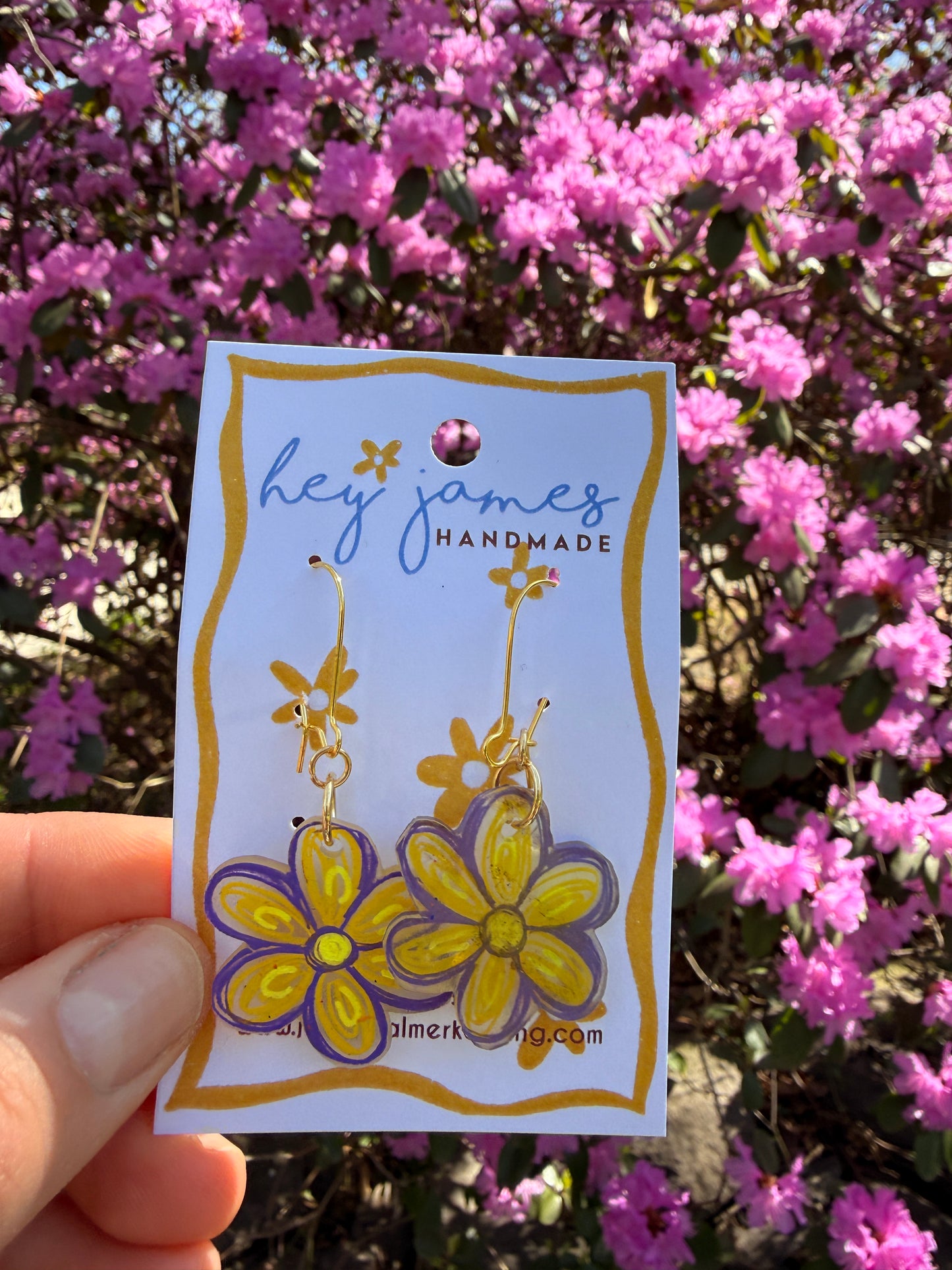 Hand holding a floral-themed yellow earrings in front of pink flowers
