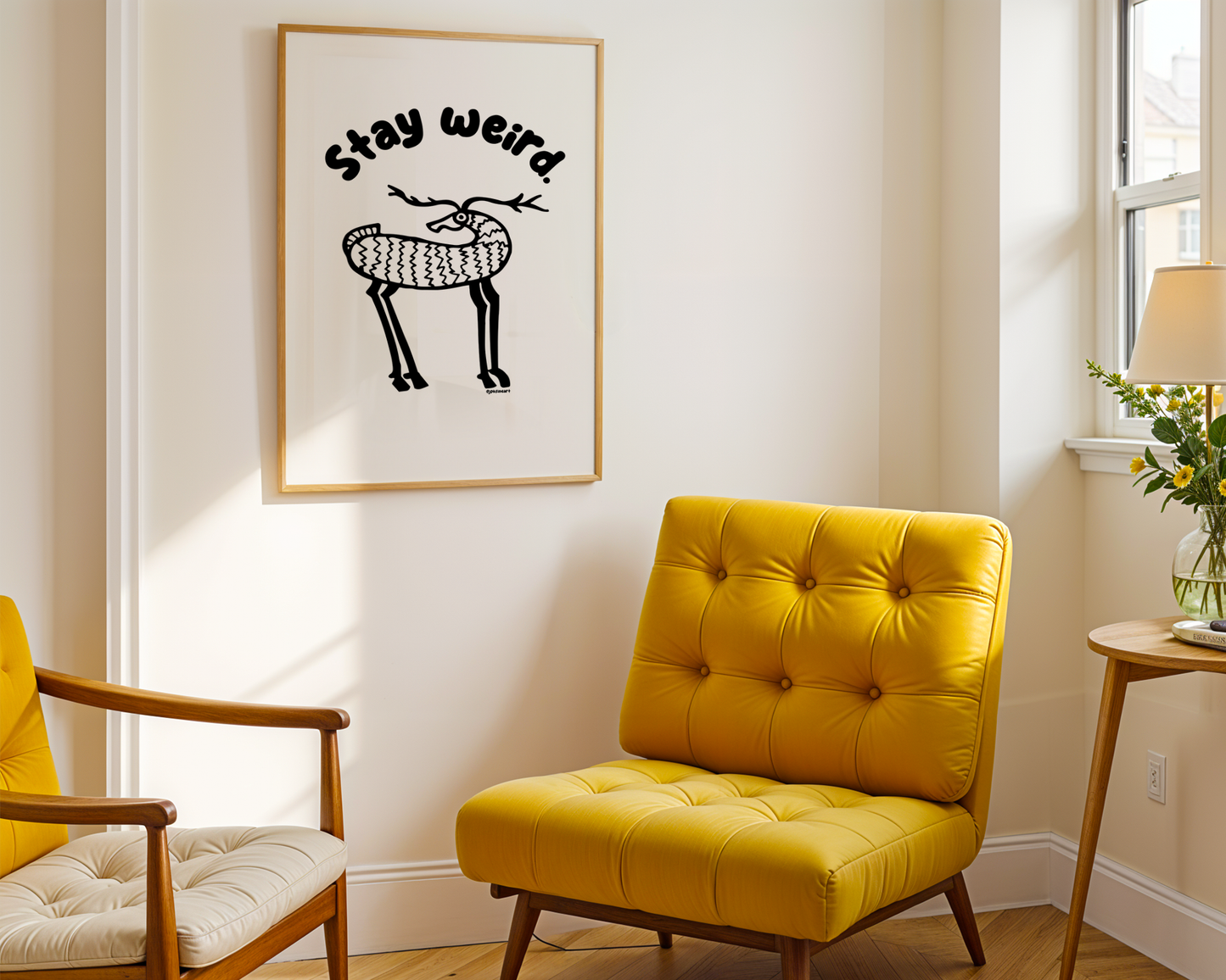 A room with a modern yellow chair shows a framed print on the wall with handwritten bubble letters spelling out “Stay Weird” Illustrated deer character in black on a white background with clean lines. Inspired by the southwest and western United States.