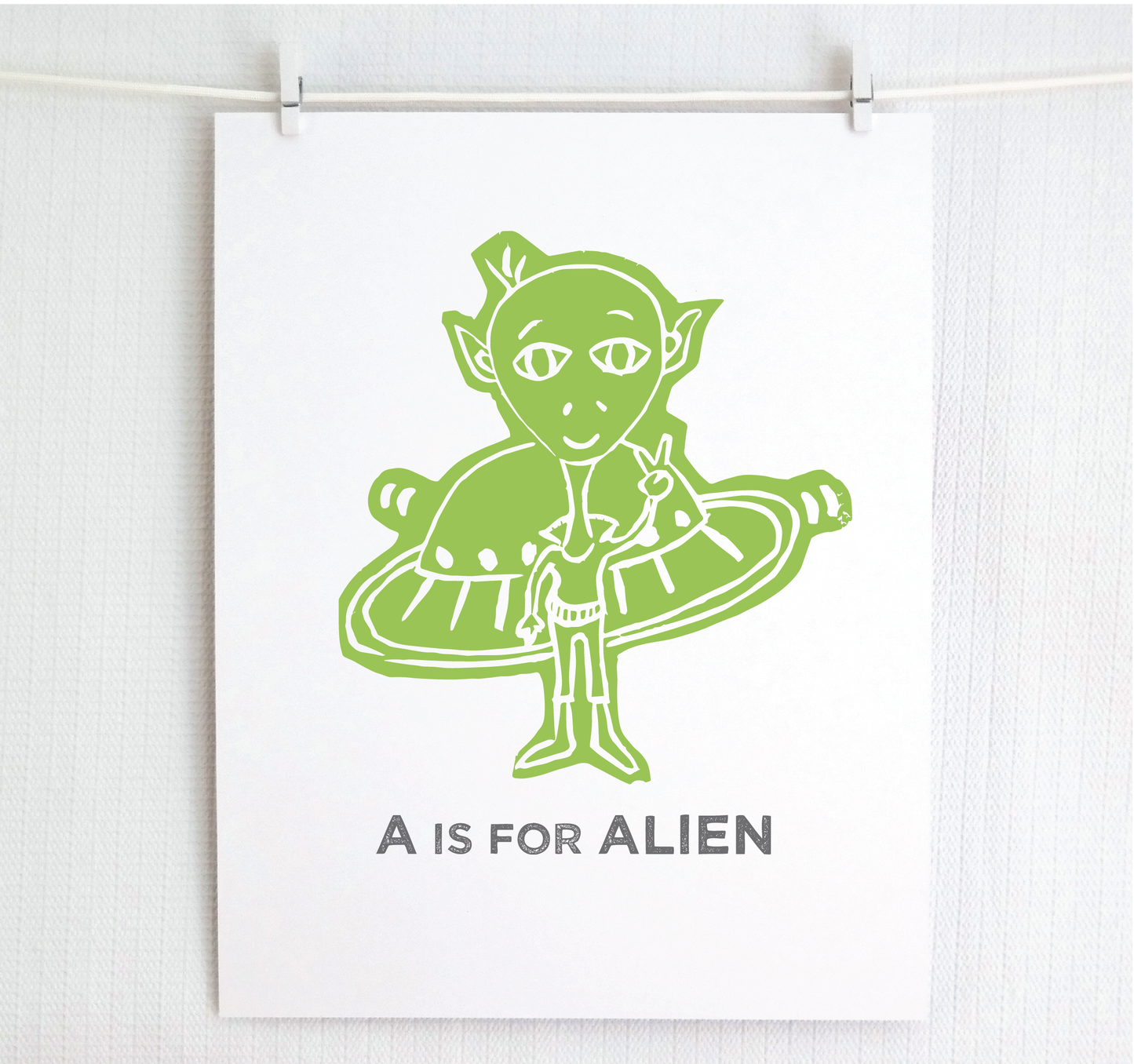 Friendly illustrated alien giving the peace sign in front of his UFO with "A is for Alien" text in a gray block font below.