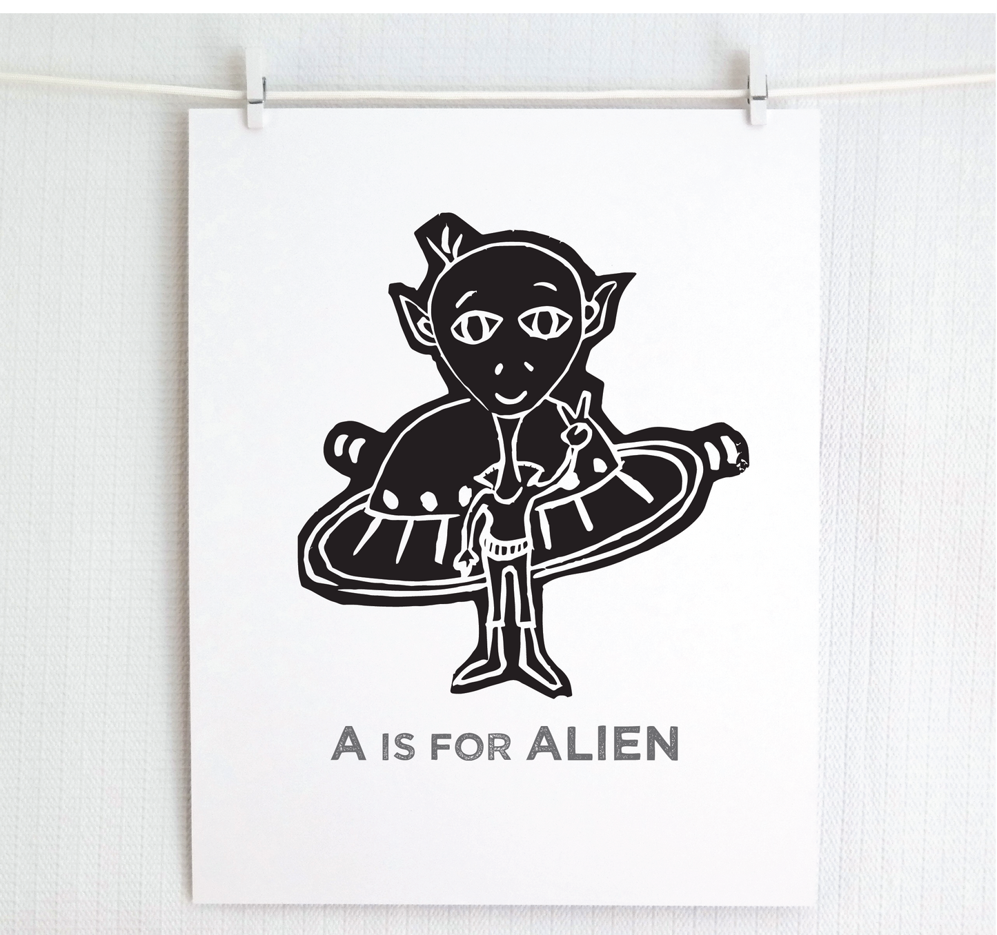 Friendly black illustrated alien giving the peace sign in front of his UFO with "A is for Alien" text in a gray block font below.