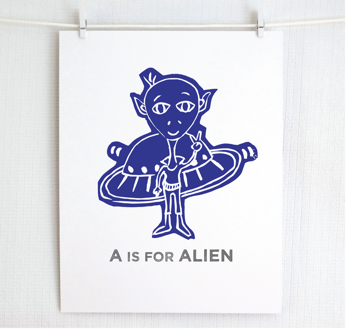 Friendly blue illustrated alien giving the peace sign in front of his UFO with "A is for Alien" text in a gray block font below.