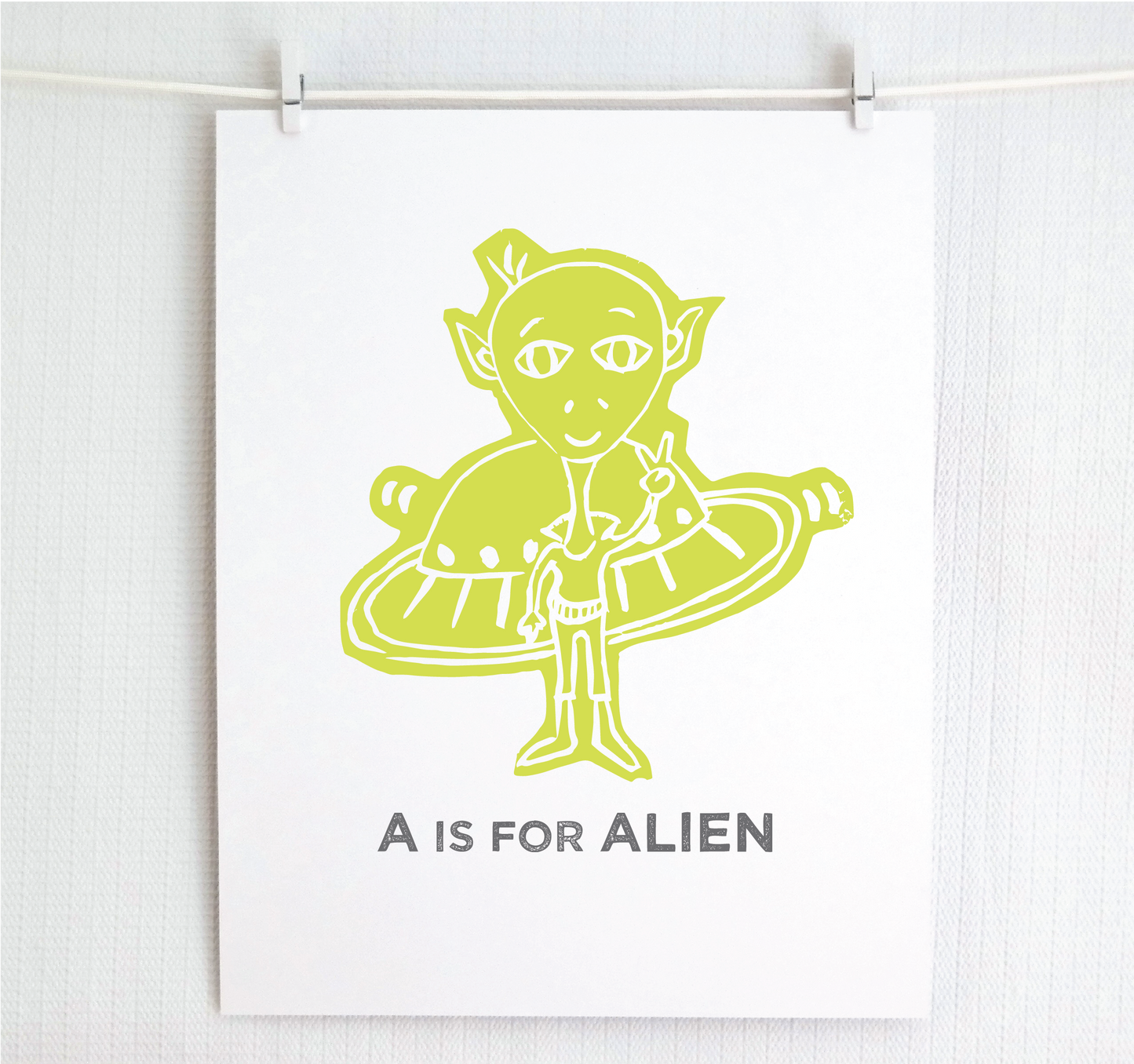 Friendly chartreuse illustrated alien giving the peace sign in front of his UFO with "A is for Alien" text in a gray block font below.