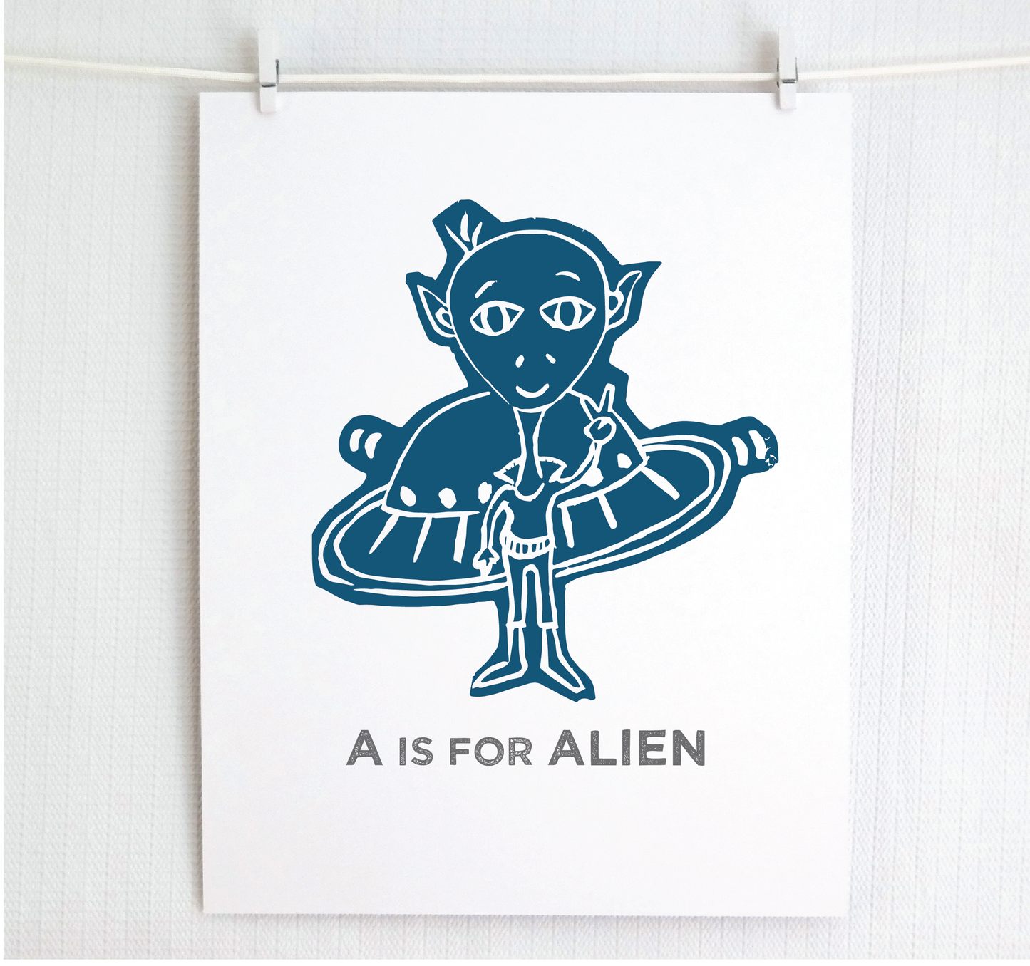 Friendly illustrated alien giving the peace sign in front of his UFO with "A is for Alien" text in a gray block font below.