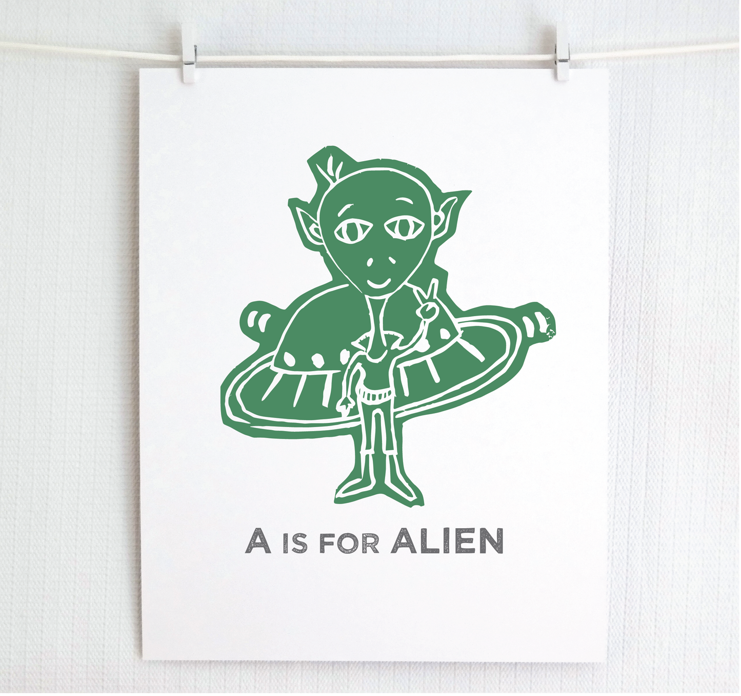 Friendly illustrated alien giving the peace sign in front of his UFO with "A is for Alien" text in a gray block font below.