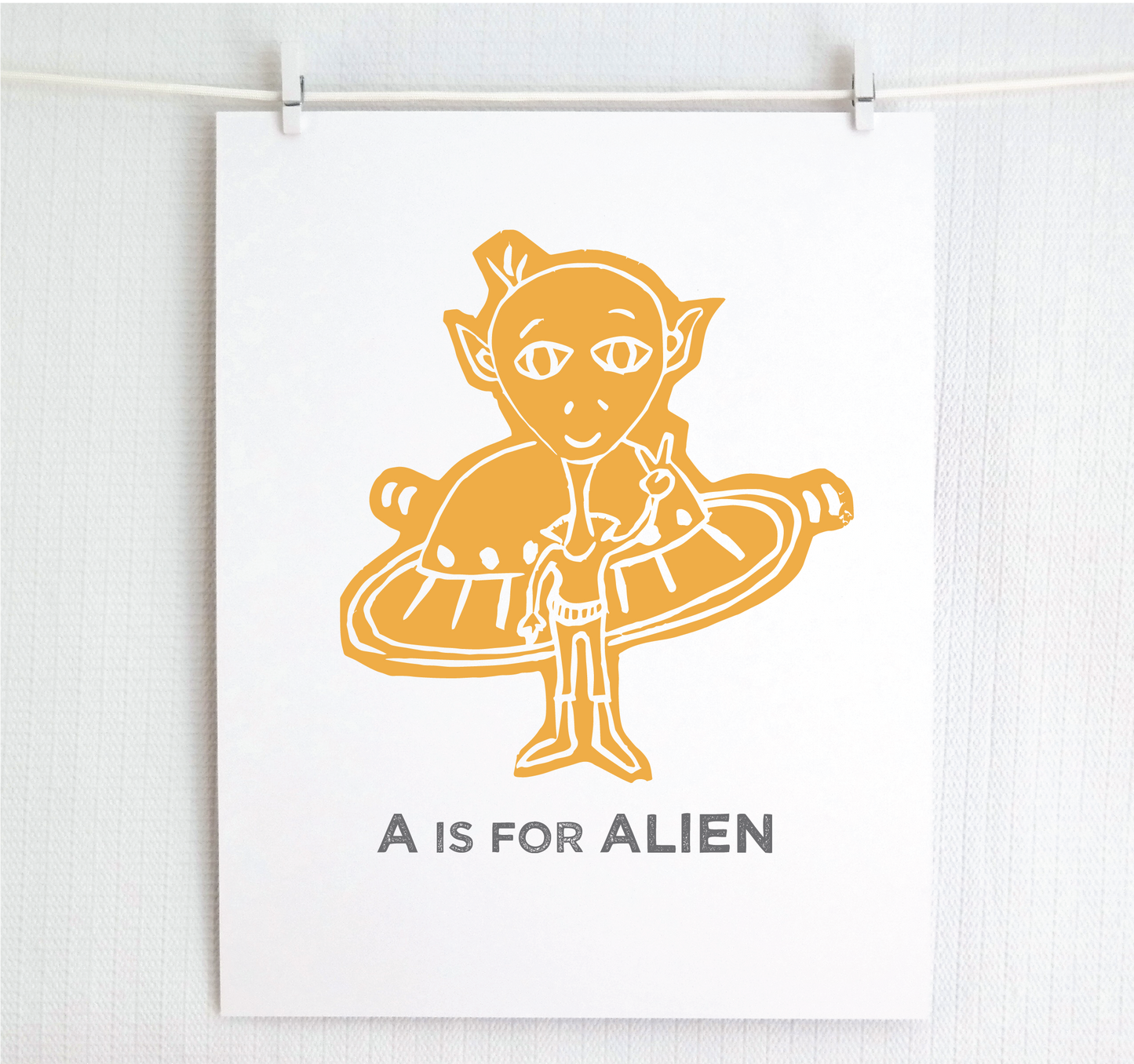 Friendly gold illustrated alien giving the peace sign in front of his UFO with "A is for Alien" text in a gray block font below.