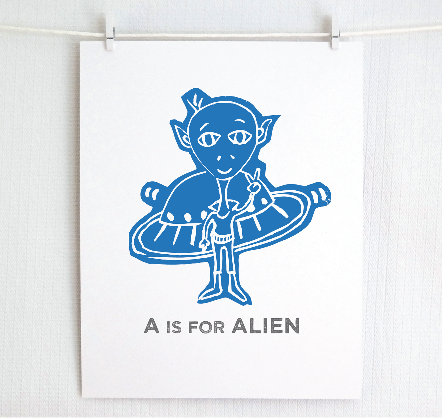 Friendly light blue illustrated alien giving the peace sign in front of his UFO with "A is for Alien" text in a gray block font below.