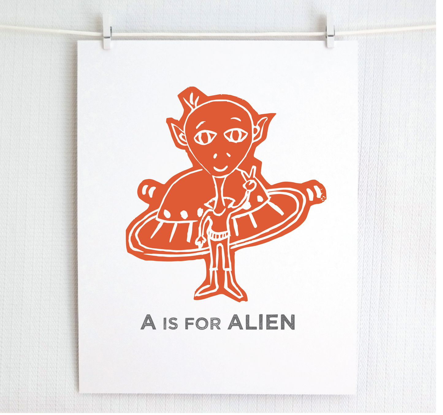 Friendly orange illustrated alien giving the peace sign in front of his UFO with "A is for Alien" text in a gray block font below.