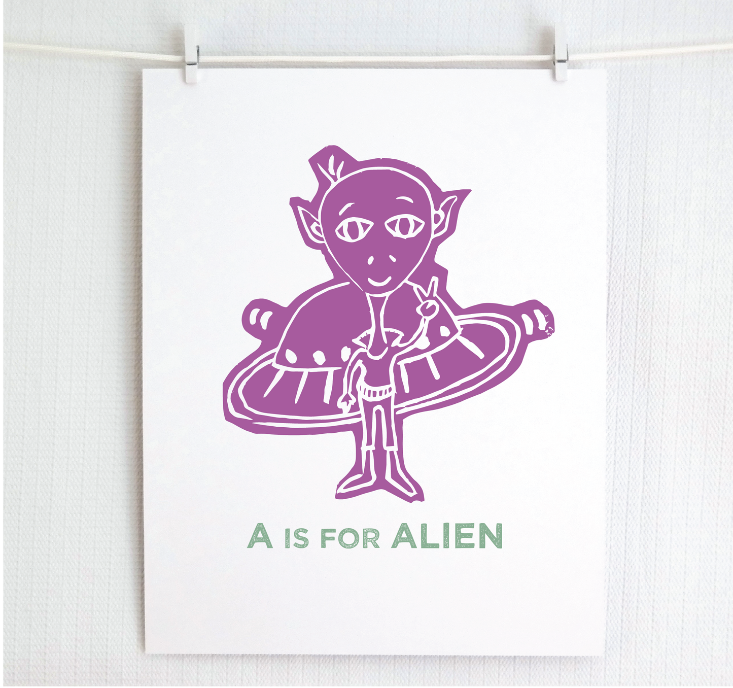 Friendly pink illustrated alien giving the peace sign in front of his UFO with "A is for Alien" text in a gray block font below.