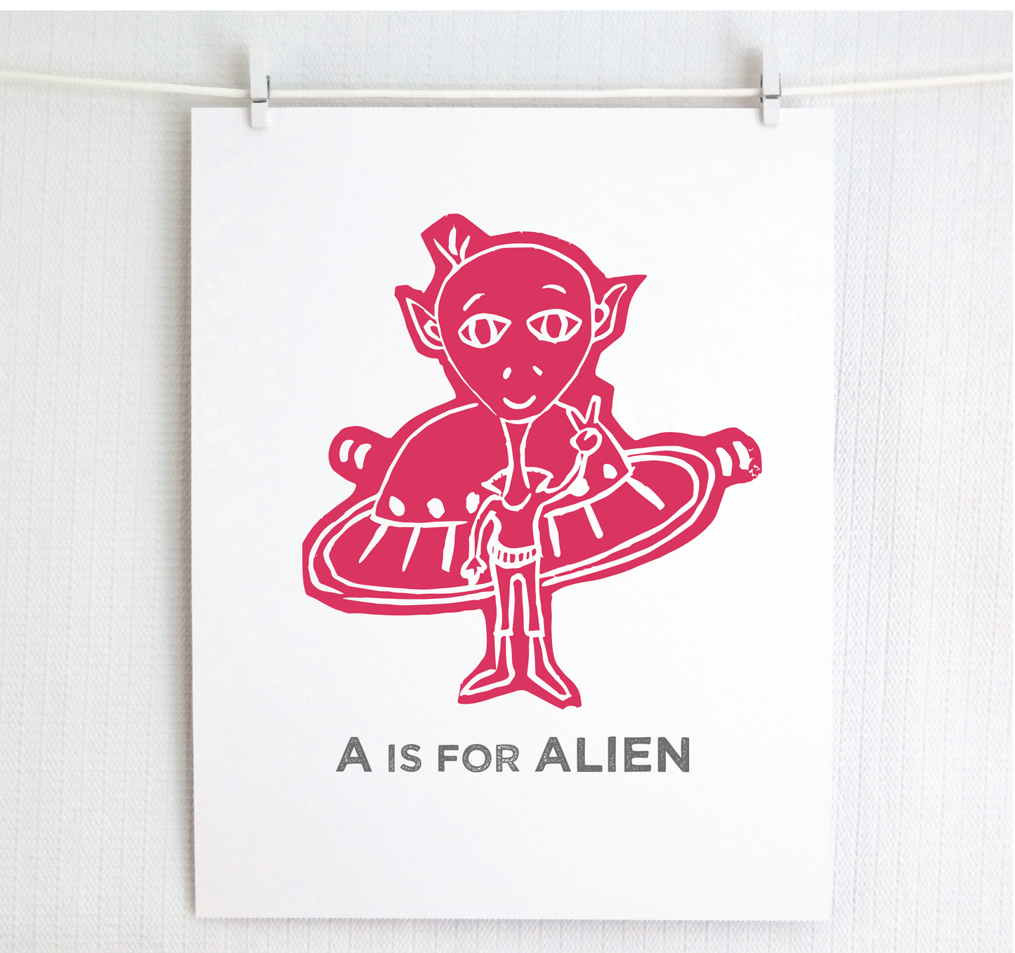 Friendly raspberry illustrated alien giving the peace sign in front of his UFO with "A is for Alien" text in a gray block font below.