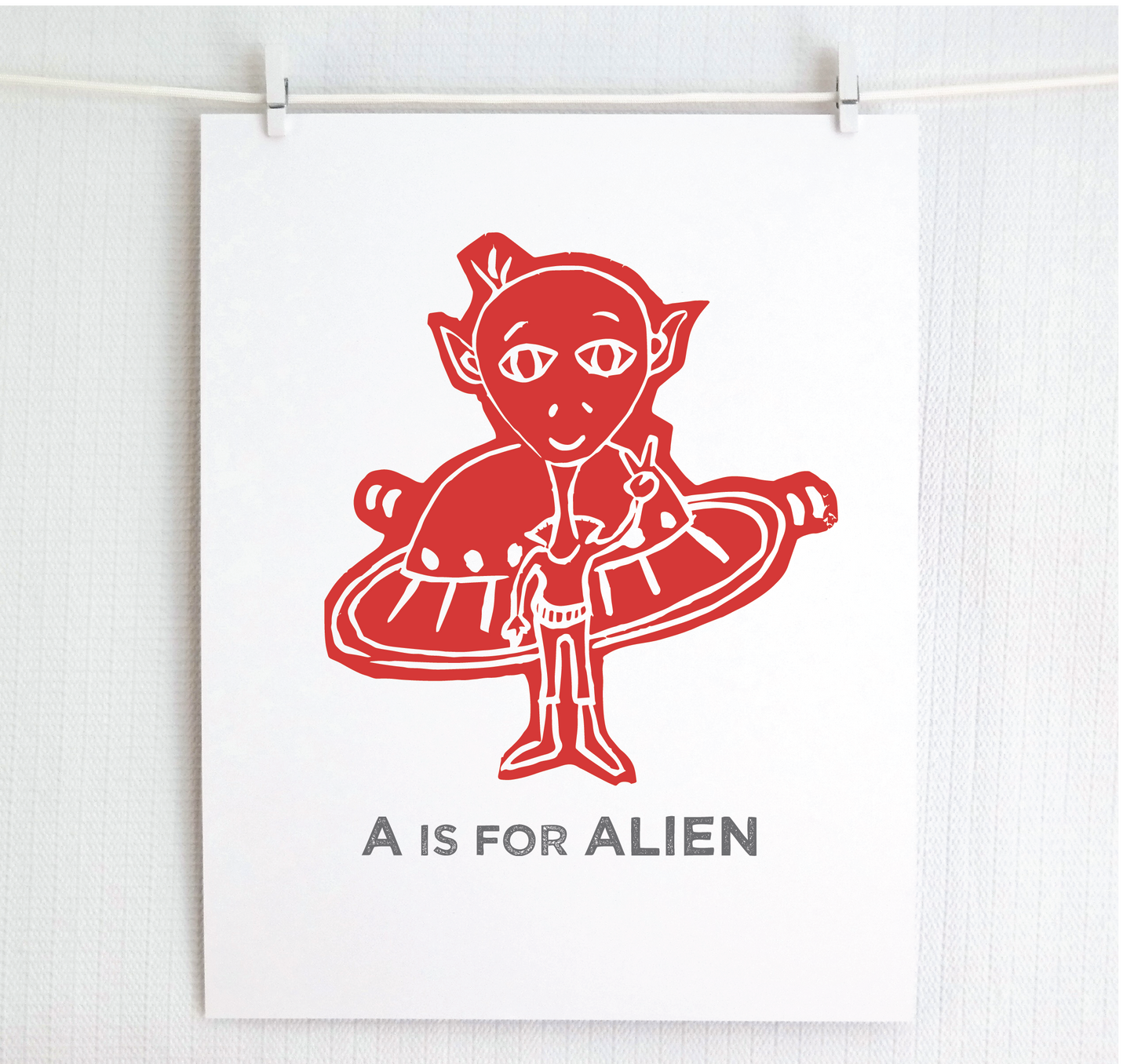 Friendly red illustrated alien giving the peace sign in front of his UFO with "A is for Alien" text in a gray block font below.