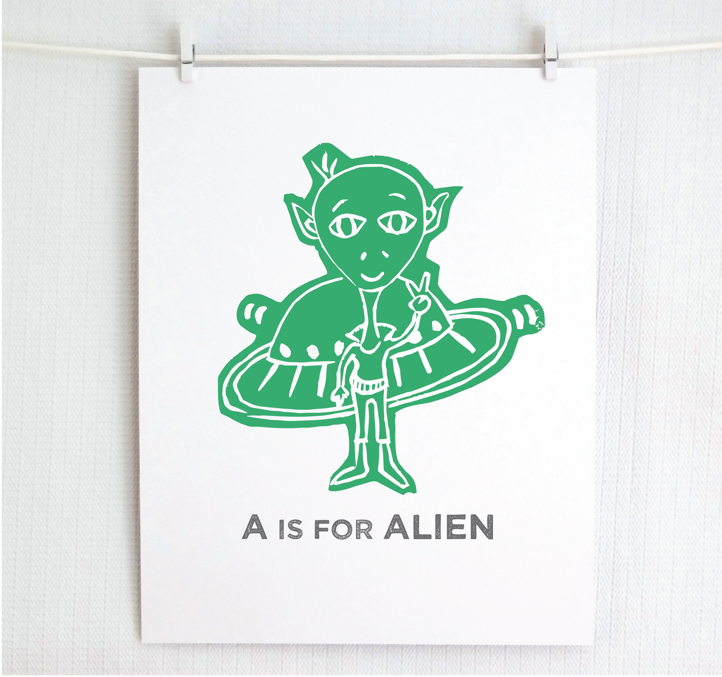 Friendly illustrated alien giving the peace sign in front of his UFO with "A is for Alien" text in a gray block font below.