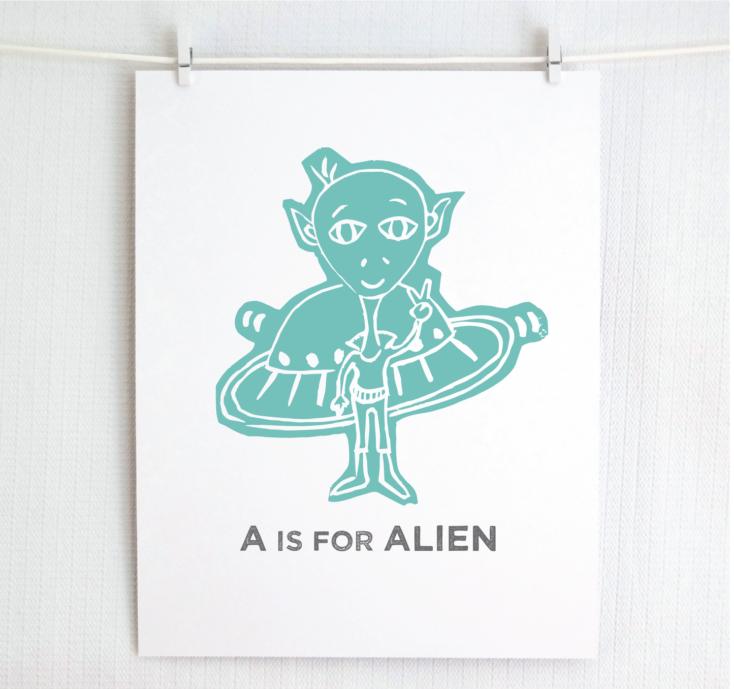 Friendly teal illustrated alien giving the peace sign in front of his UFO with "A is for Alien" text in a gray block font below.