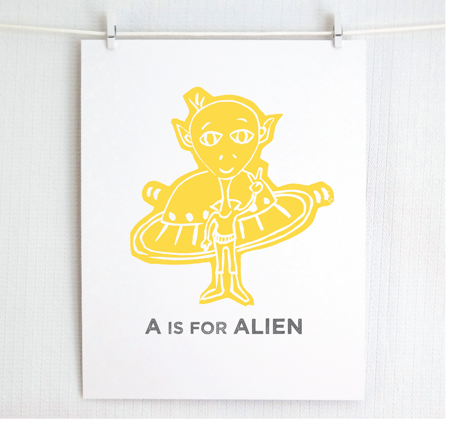 Friendly yellow illustrated alien giving the peace sign in front of his UFO with "A is for Alien" text in a gray block font below.