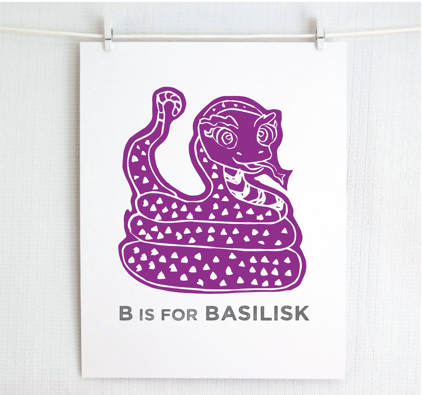 Friendly illustrated basilisk giving a slithery smile above “B is for Basilisk” text in a gray block font below.