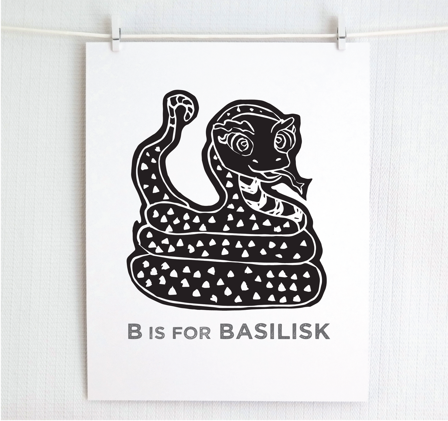 Friendly illustrated basilisk giving a slithery smile above “B is for Basilisk” text in a gray block font below.