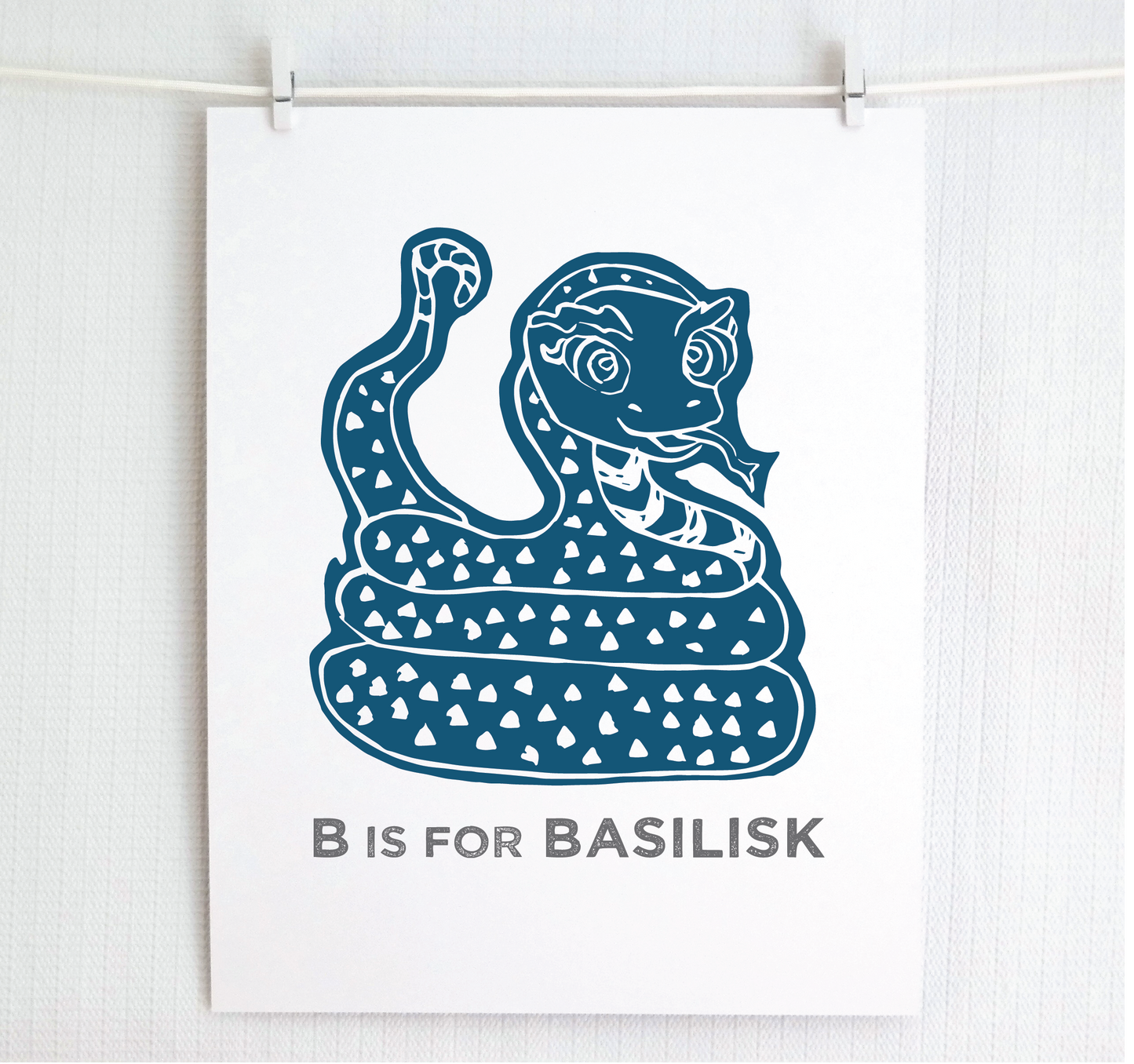 Friendly illustrated basilisk giving a slithery smile above “B is for Basilisk” text in a gray block font below.