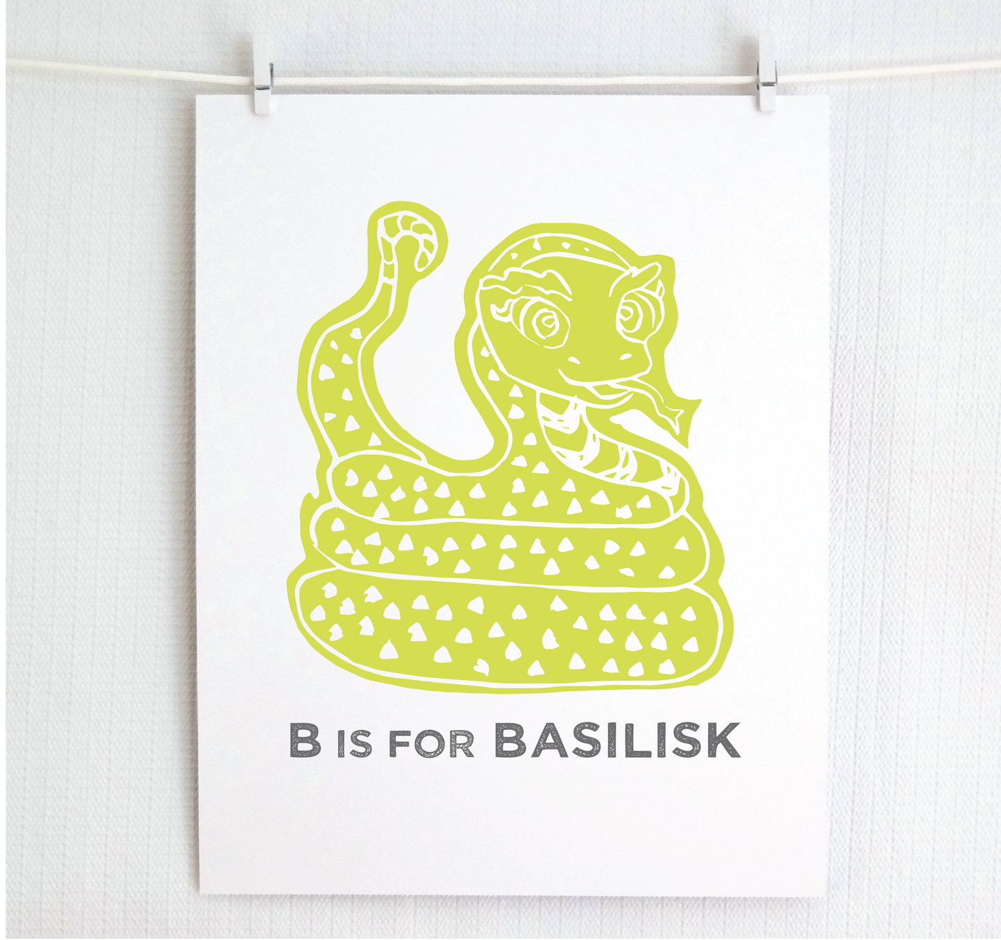 Friendly illustrated basilisk giving a slithery smile above “B is for Basilisk” text in a gray block font below.