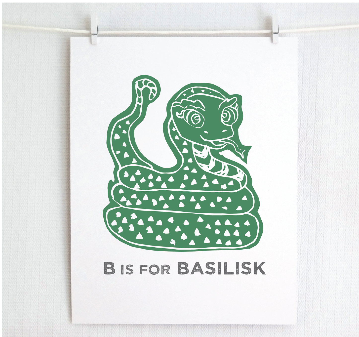 Friendly illustrated basilisk giving a slithery smile above “B is for Basilisk” text in a gray block font below.