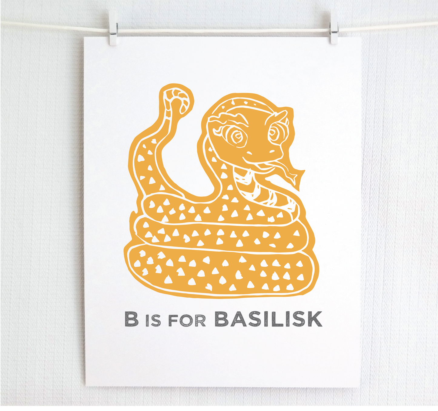Friendly illustrated basilisk giving a slithery smile above “B is for Basilisk” text in a gray block font below.