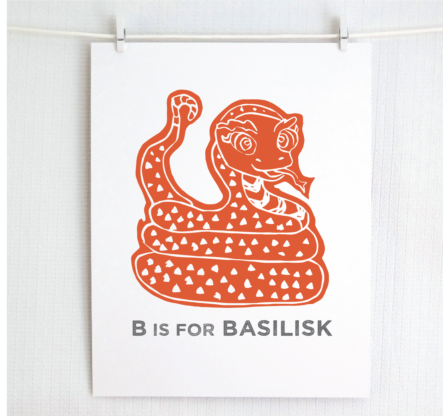 Friendly illustrated basilisk giving a slithery smile above “B is for Basilisk” text in a gray block font below.