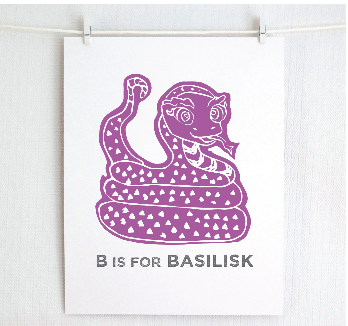 Friendly illustrated basilisk giving a slithery smile above “B is for Basilisk” text in a gray block font below.