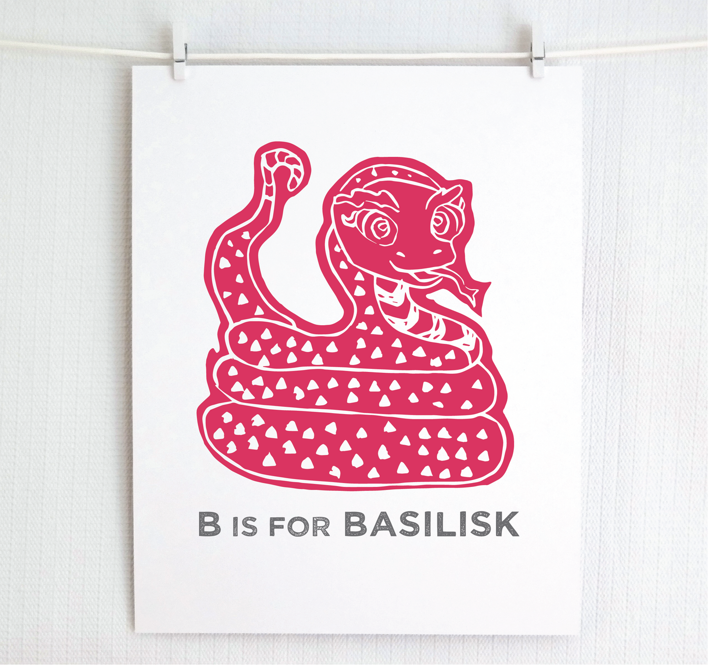 Friendly illustrated basilisk giving a slithery smile above “B is for Basilisk” text in a gray block font below.