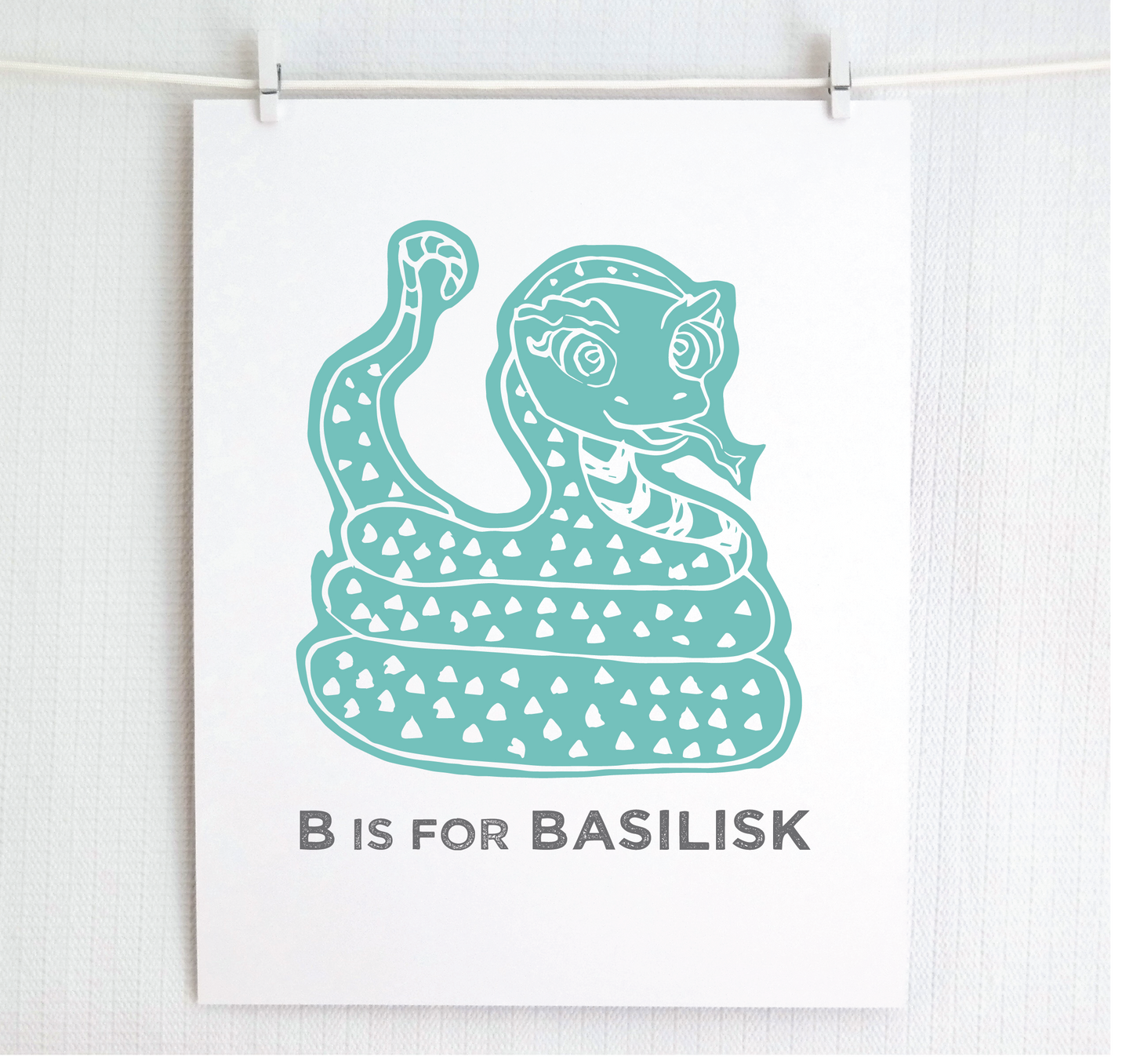 Friendly illustrated basilisk giving a slithery smile above “B is for Basilisk” text in a gray block font below.
