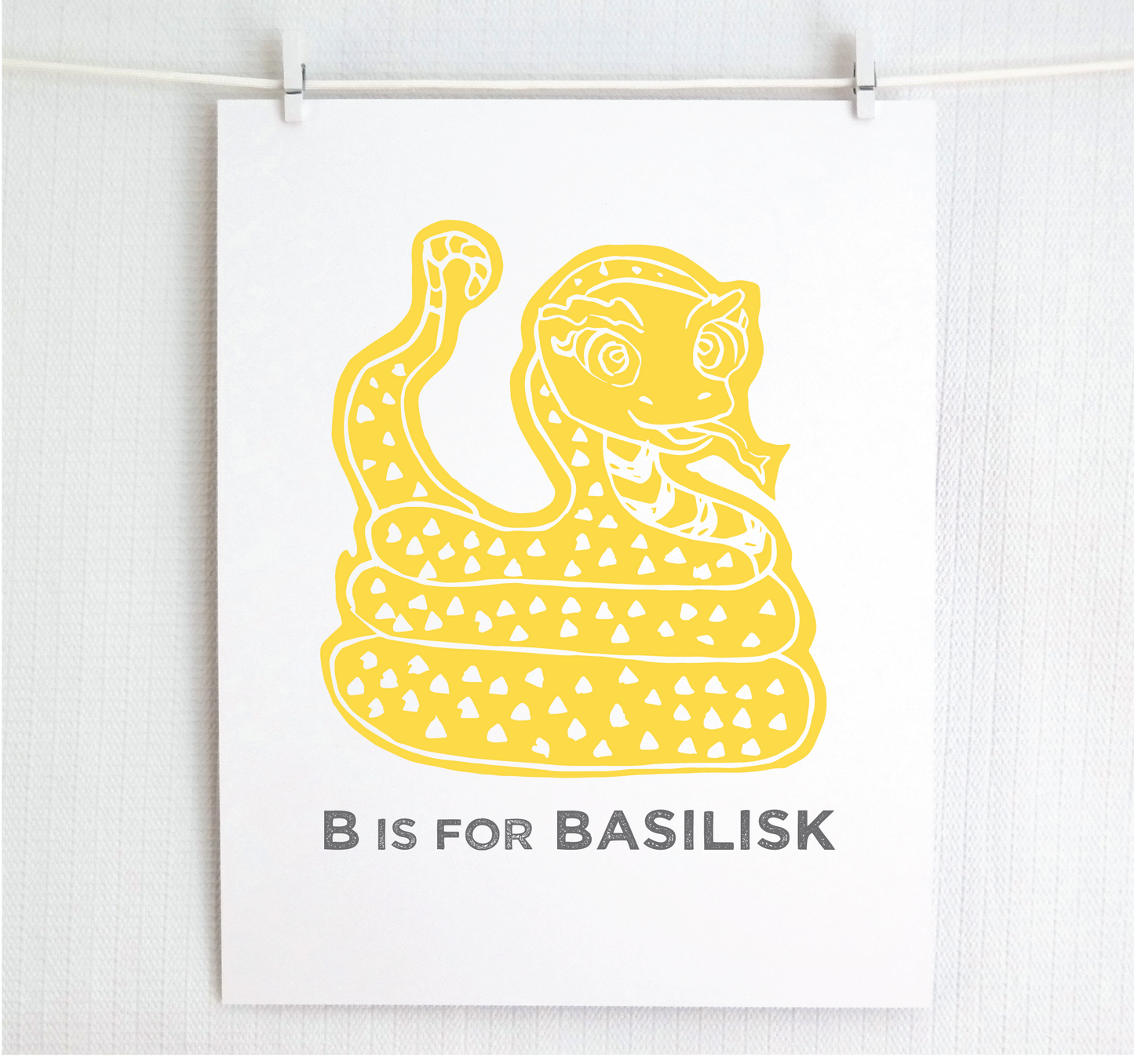 Friendly illustrated basilisk giving a slithery smile above “B is for Basilisk” text in a gray block font below.