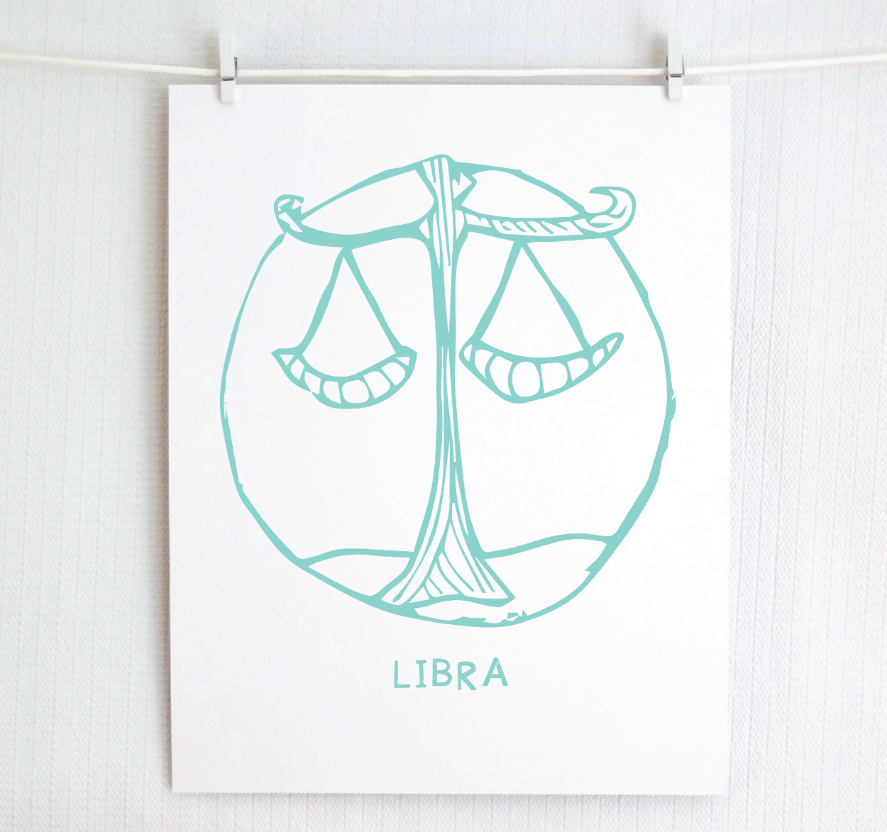 An illustration of the LIBRA Zodiac sign, printed on paper with the word 'LIBRA’ at the bottom in light teal.