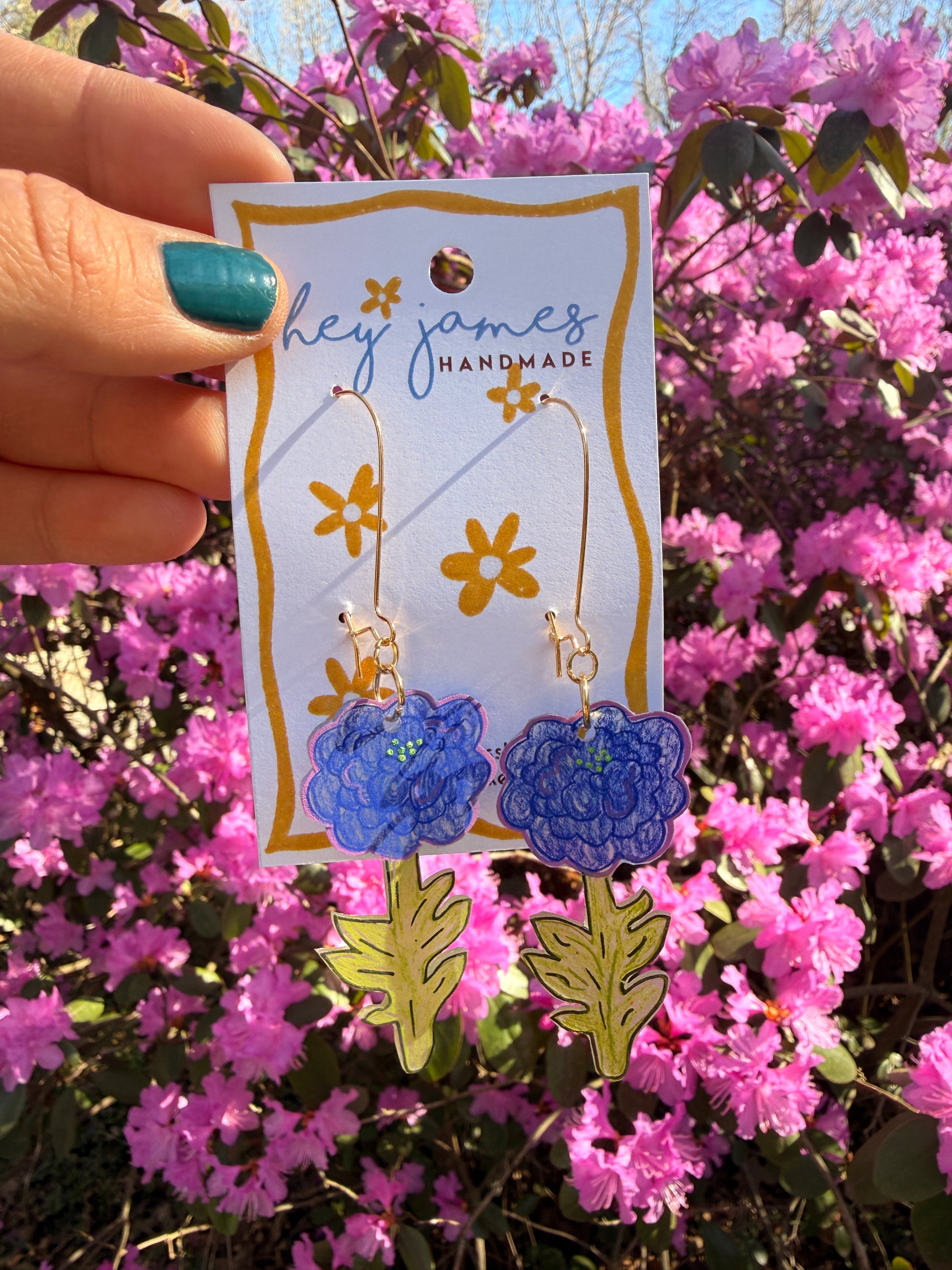 Handmade earrings with floral design on a card held in front of pink flowers