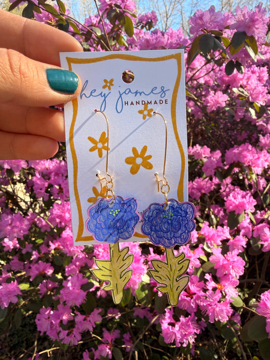 Handmade earrings with floral design on a card held in front of pink flowers