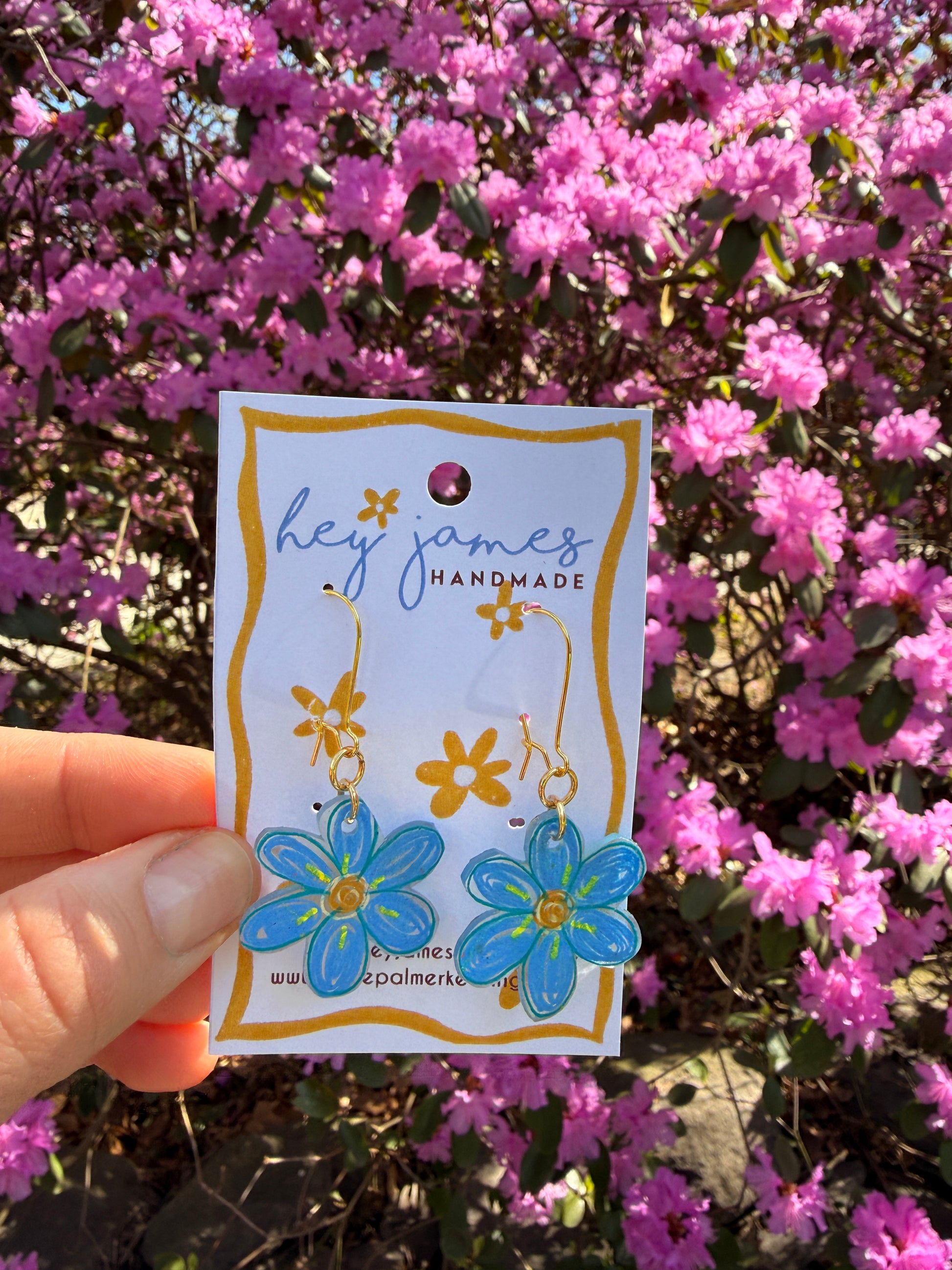 Hand holding a floral-themed blue earrings in front of pink flowers