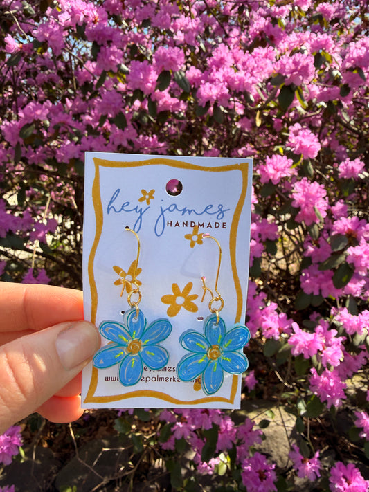Hand holding a floral-themed blue earrings in front of pink flowers