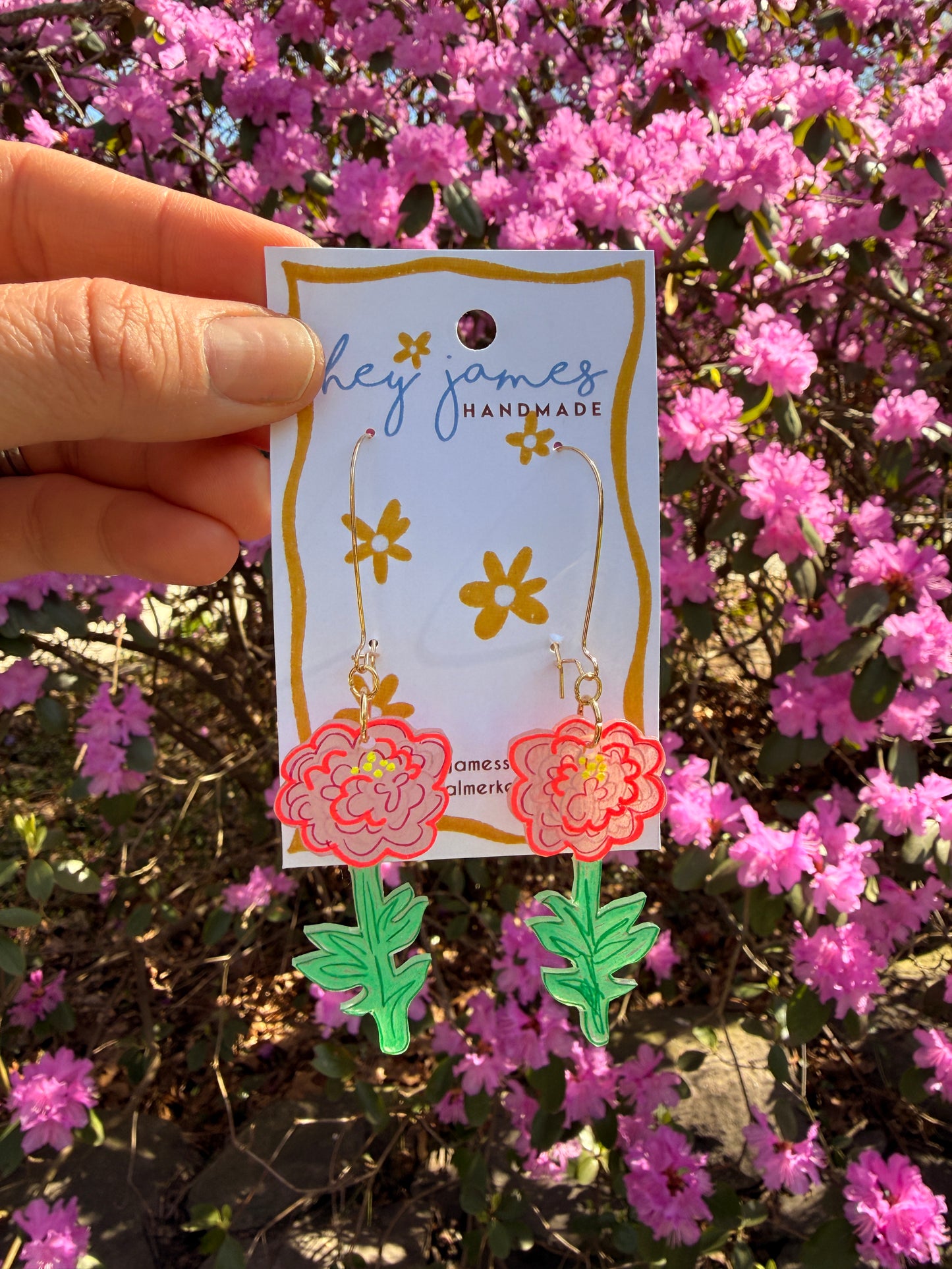 Handmade earrings with floral design held against a background of pink flowers