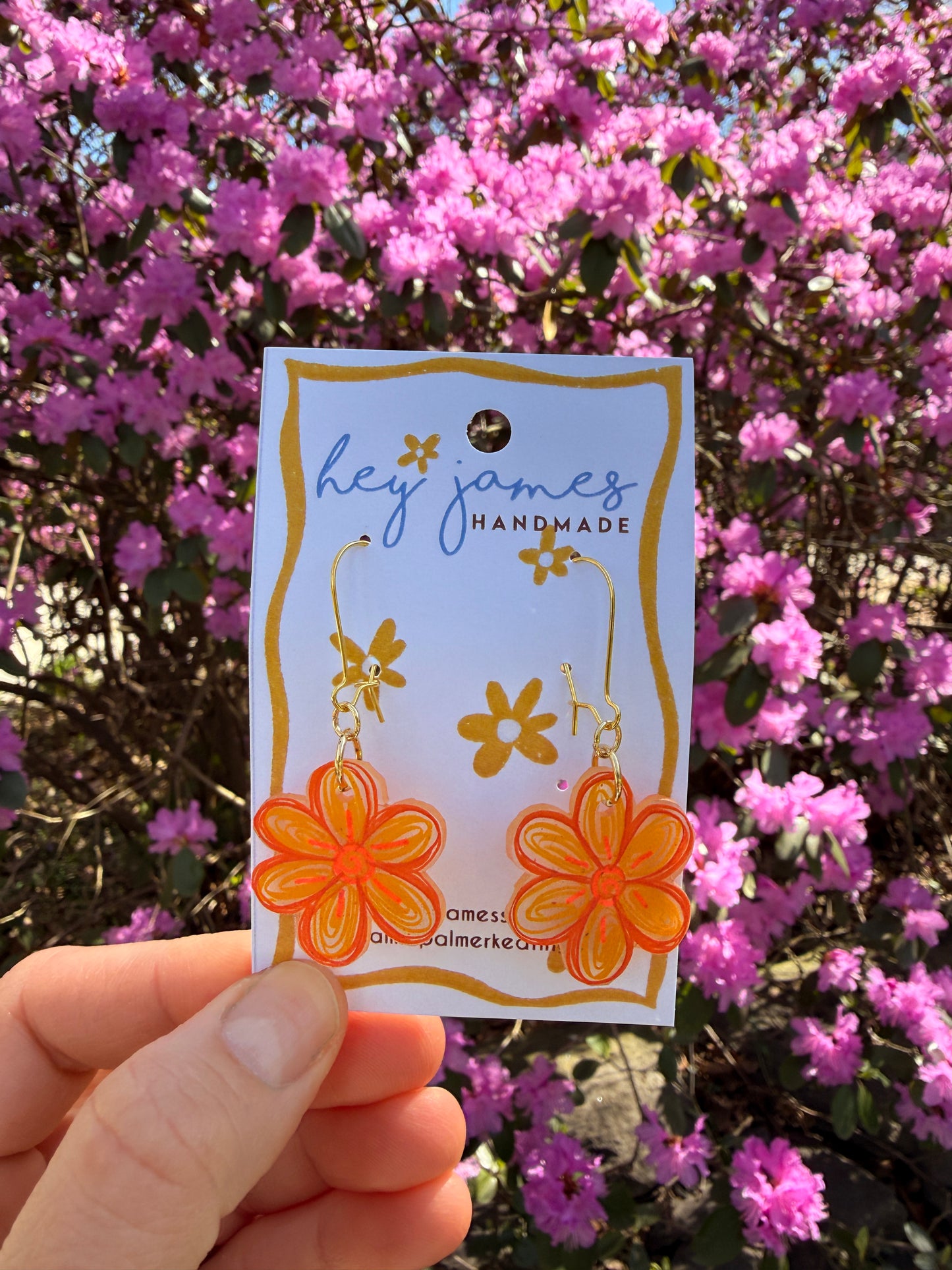 Hand holding a card with floral earrings and 'hey james handmade' text in front of pink flowers