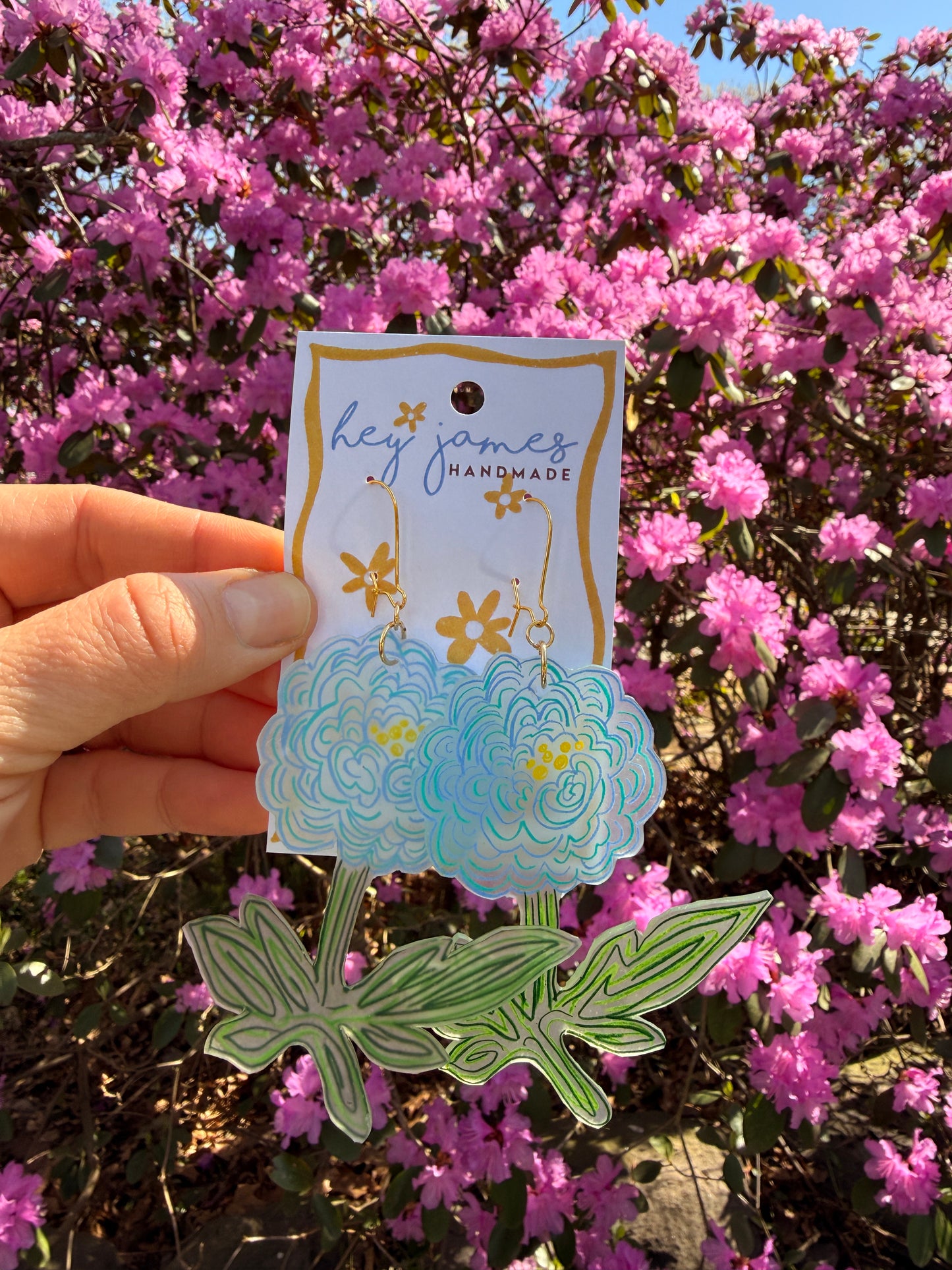 Hand holding decorative blue flower earrings with floral designs against a pink flowering bush.