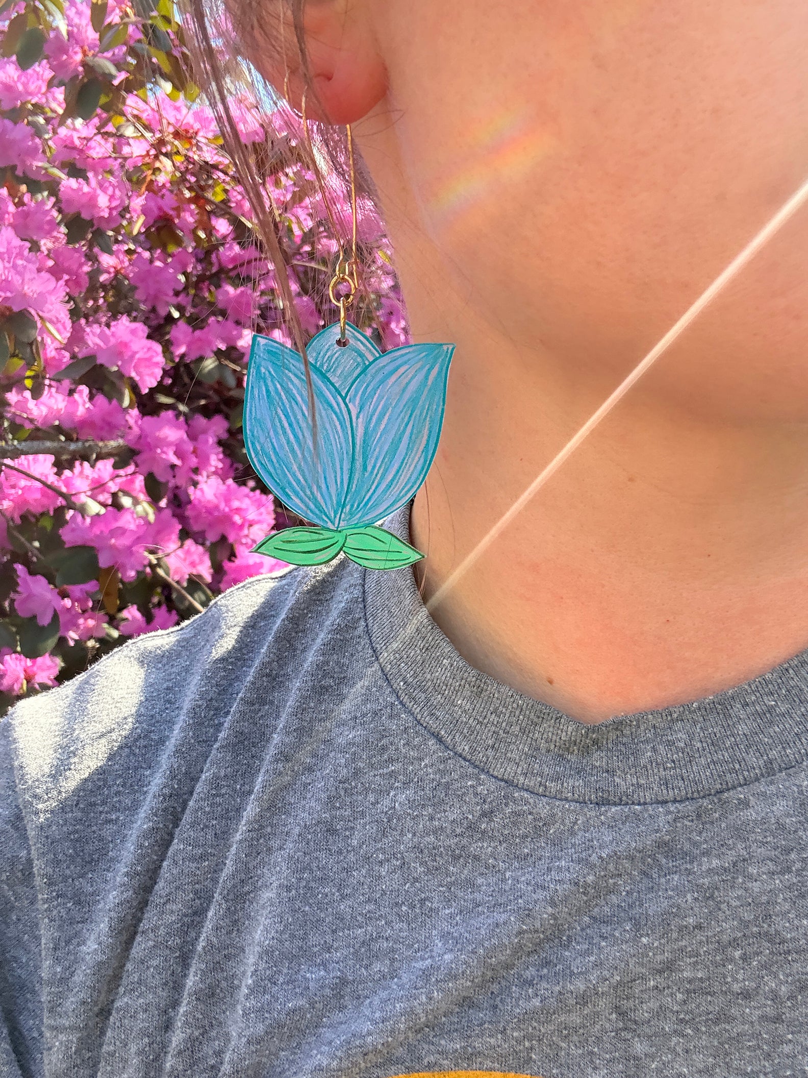 Person wearing a blue and green leaf-shaped earring with pink flowers in the background