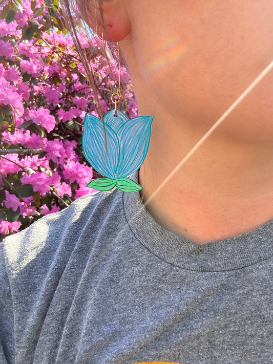 Person wearing a blue and green leaf-shaped earring with pink flowers in the background