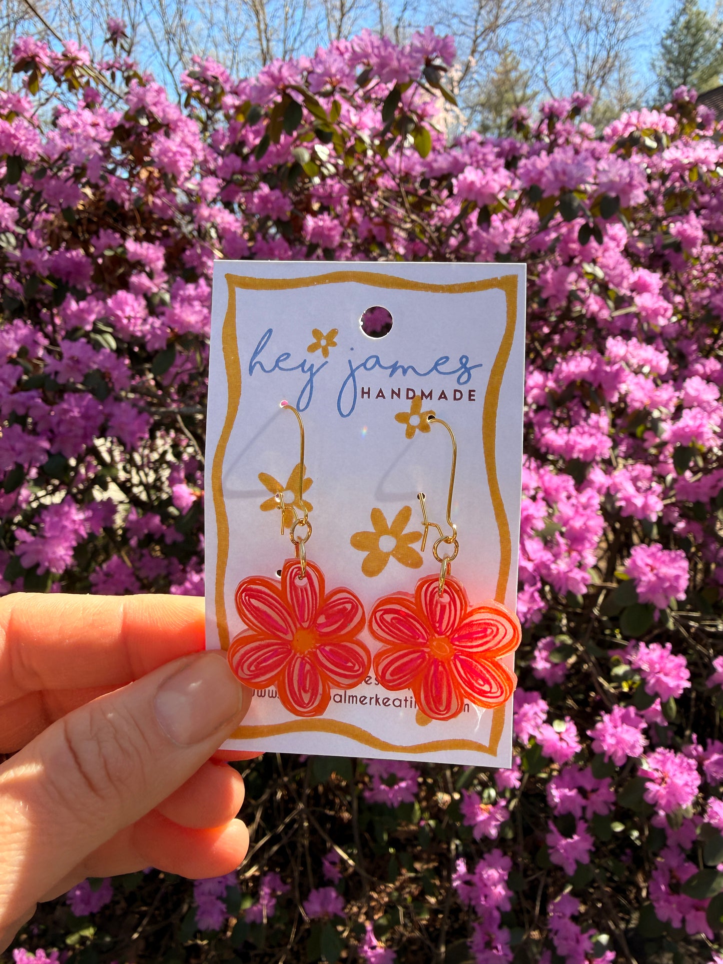 Hand holding bright pink floral earrings against a pink flowering bush.