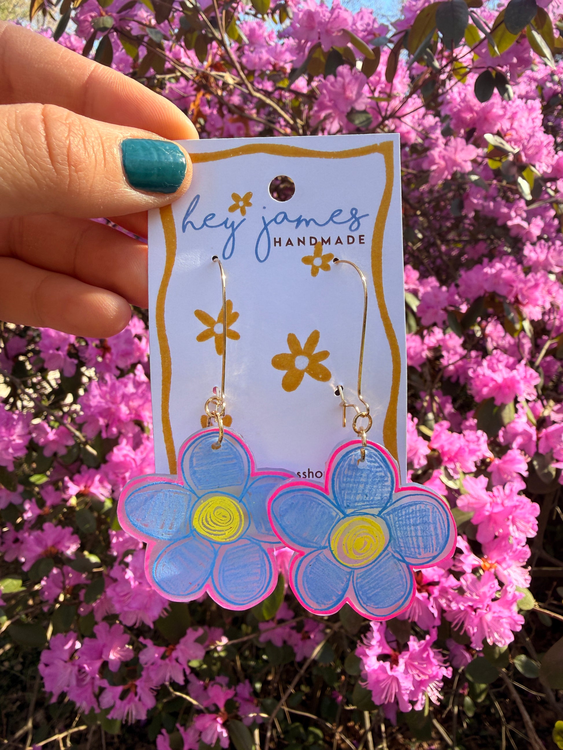 Flower-shaped earrings on a 'hey james' card held in front of pink flowers