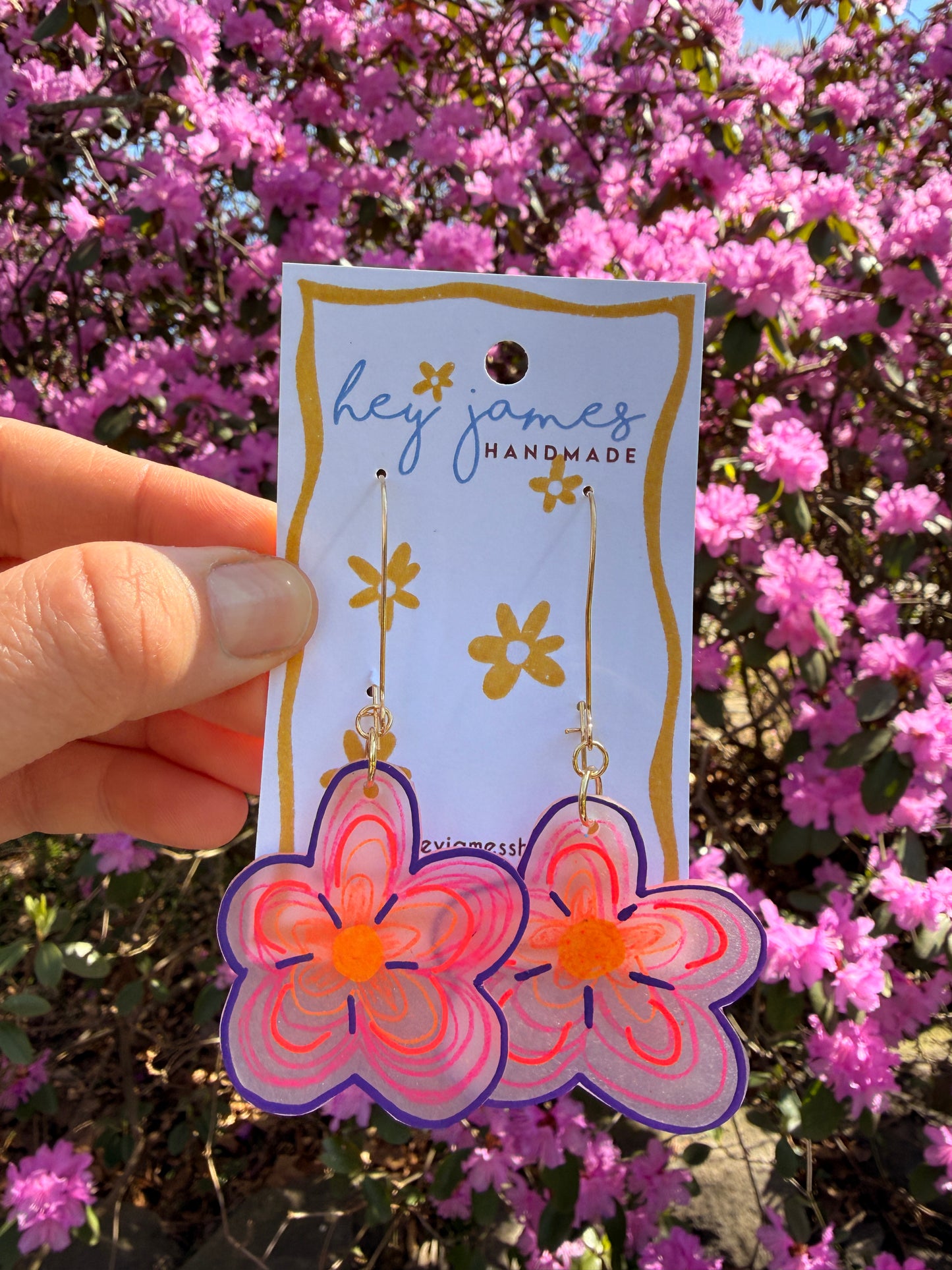 Flower-shaped earrings on a card held in front of pink flowers