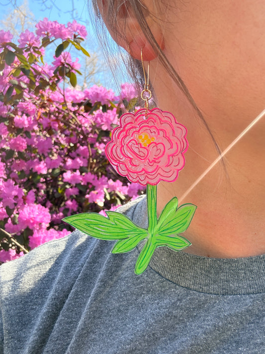 Close-up of a pink flower earring with green leaves worn by a person against a blurred natural background.
