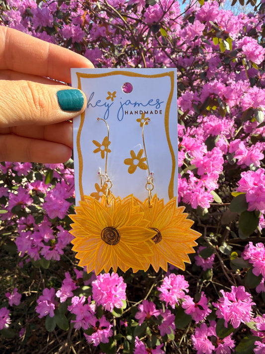 Hand holding a card with sunflower earrings against a background of pink flowers