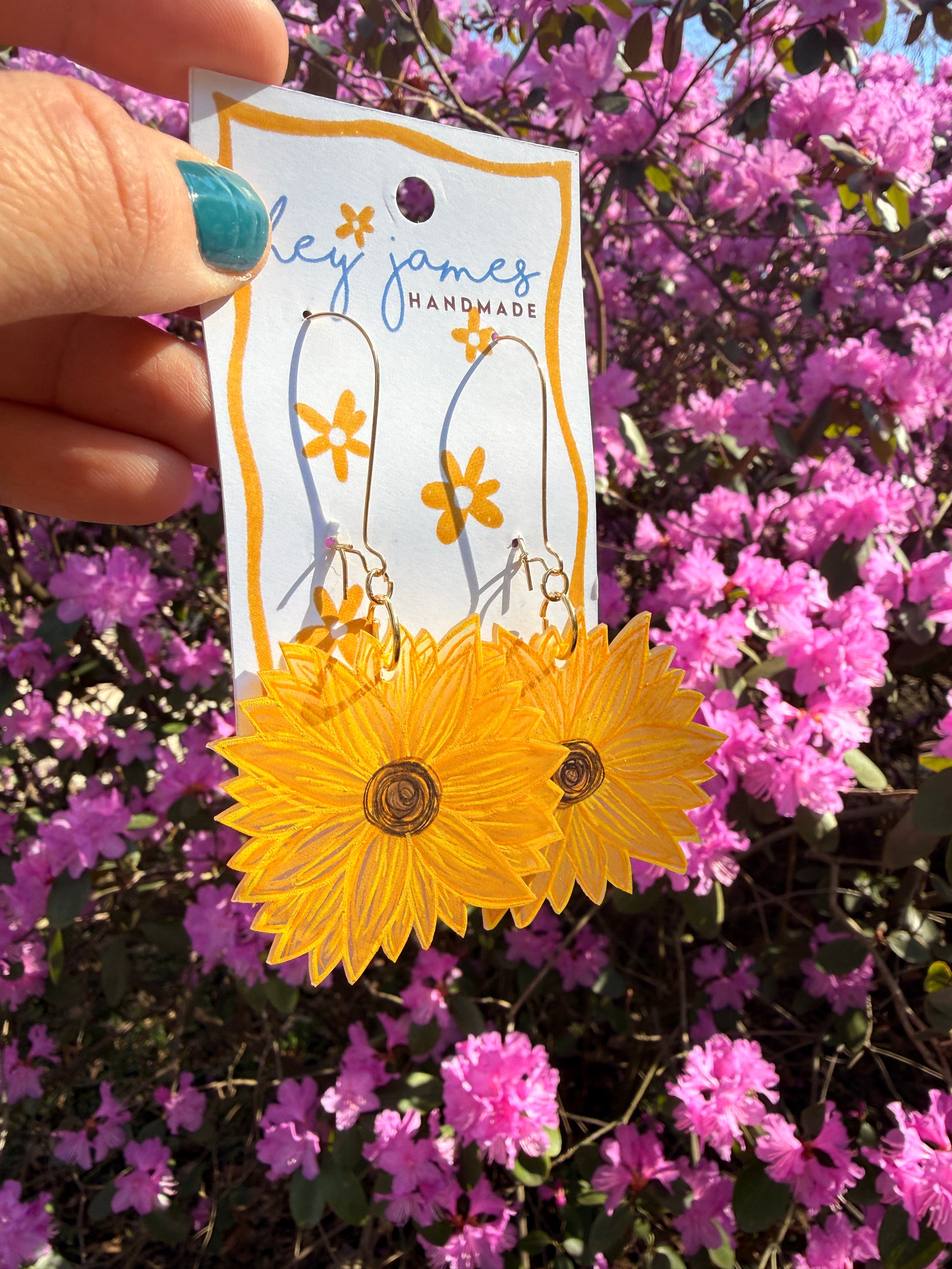 Yellow sunflower-shaped earrings held against a background of pink flowers