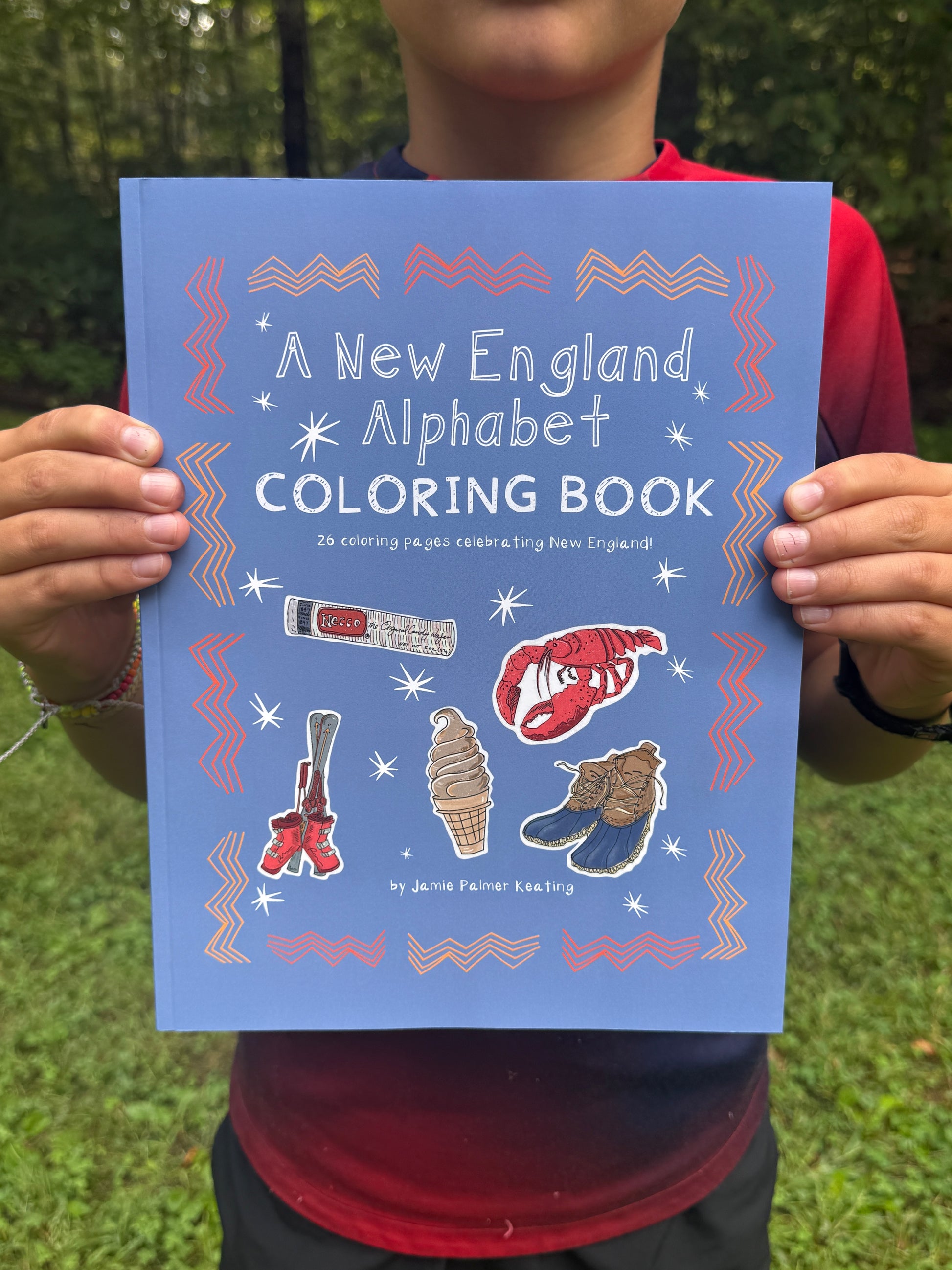 Person holding a 'New England Alphabet Coloring Book' with illustrations on a grassy background