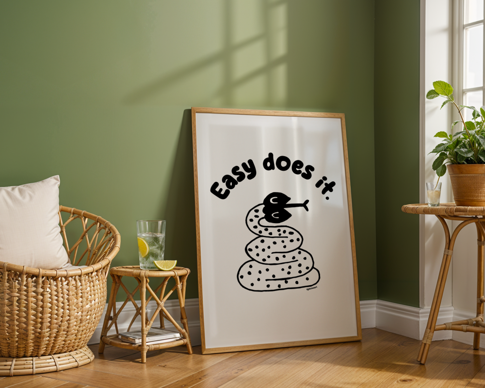 Green walled room with large frame on the floor showing print with handwritten thin letters spelling out “Easy Does It.” Illustrated snake character in black on a white background with clean lines. Inspired by the western United States.
