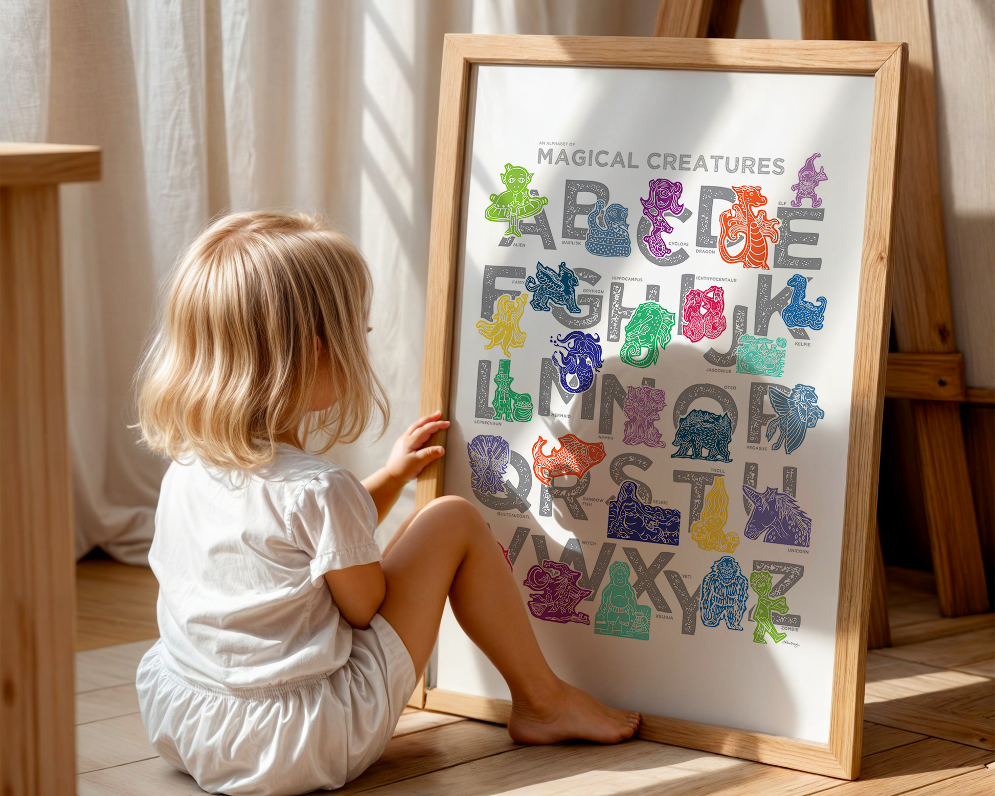 A little kid sits on the floor and admires a colorful illustrated magical creature alphabet with a creature for every letter of the alphabet printed on a white background. Chunky illustrated letters in gray sit behind each character. Colors are jewel-toned and include pinks and purples, blues and greens, yellows and reds.
