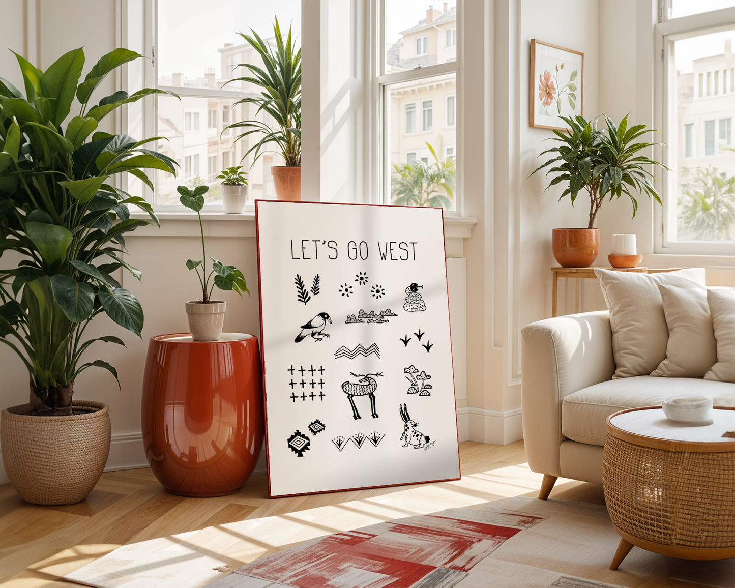 Handwritten thin letters spelling out “Let’s Go West” framed print on a living room floor surrounded by plants and neutral colors. Illustrated characters in black on a. white background with clean lines, include snake, crow, deer and rabbit with flora and fauna. Inspired by the southwest and western United States.
